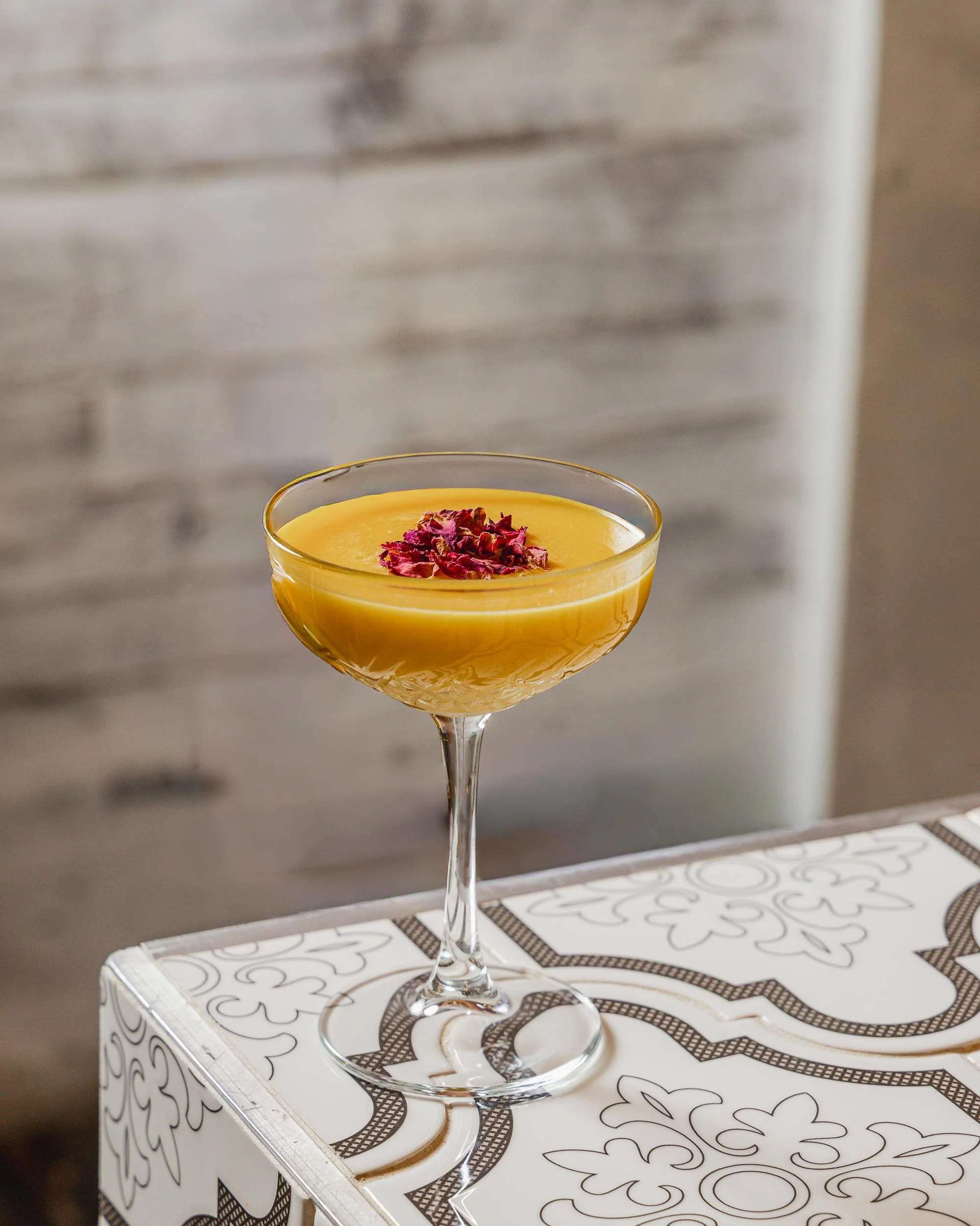 A yellow cocktail garnished with dried rose petals in a crystal glass on a decorative white and black patterned table.