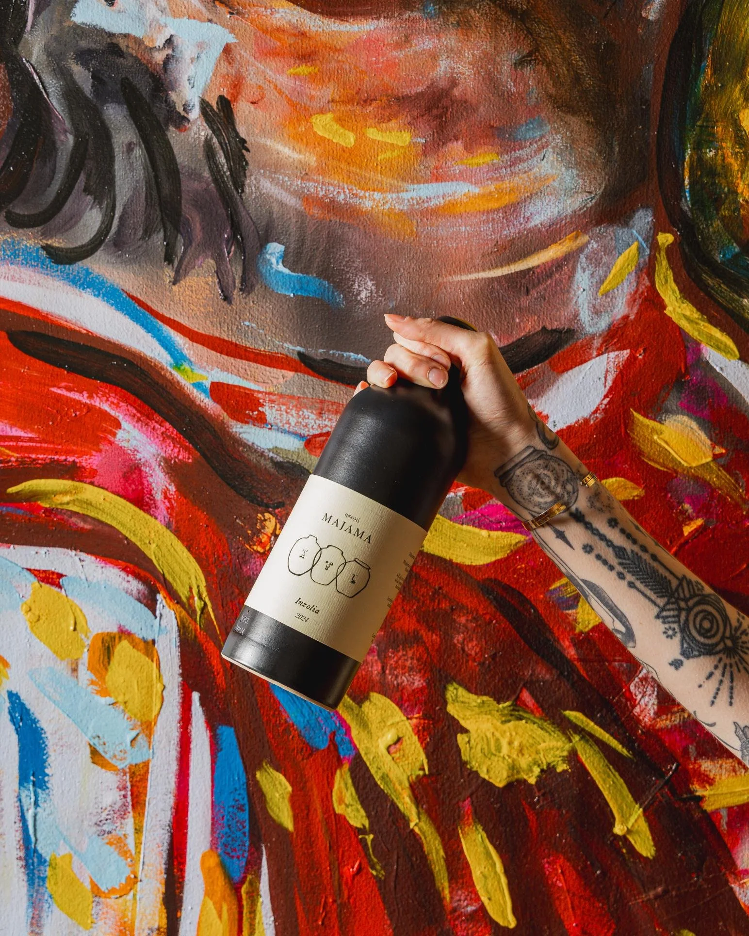A person with tattoos on their arm holding a black and white bottle in front of an abstract, colorful painting.