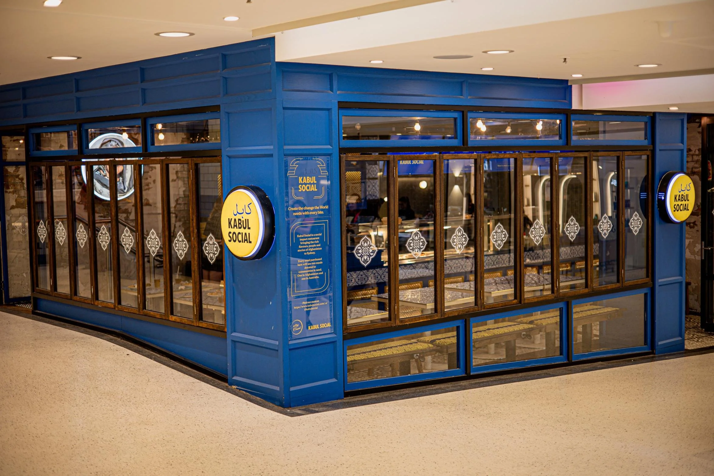 A restaurant named Kabul Social with blue exterior walls and large glass windows, decorated with white geometric patterns. The sign displays the restaurant's name in English and Arabic.