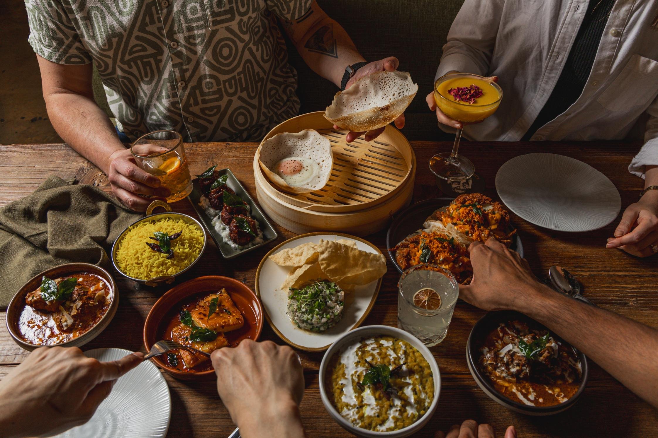 A group of people sharing an Indian meal with various dishes, including rice, curry, bread, and drinks, on a wooden table.