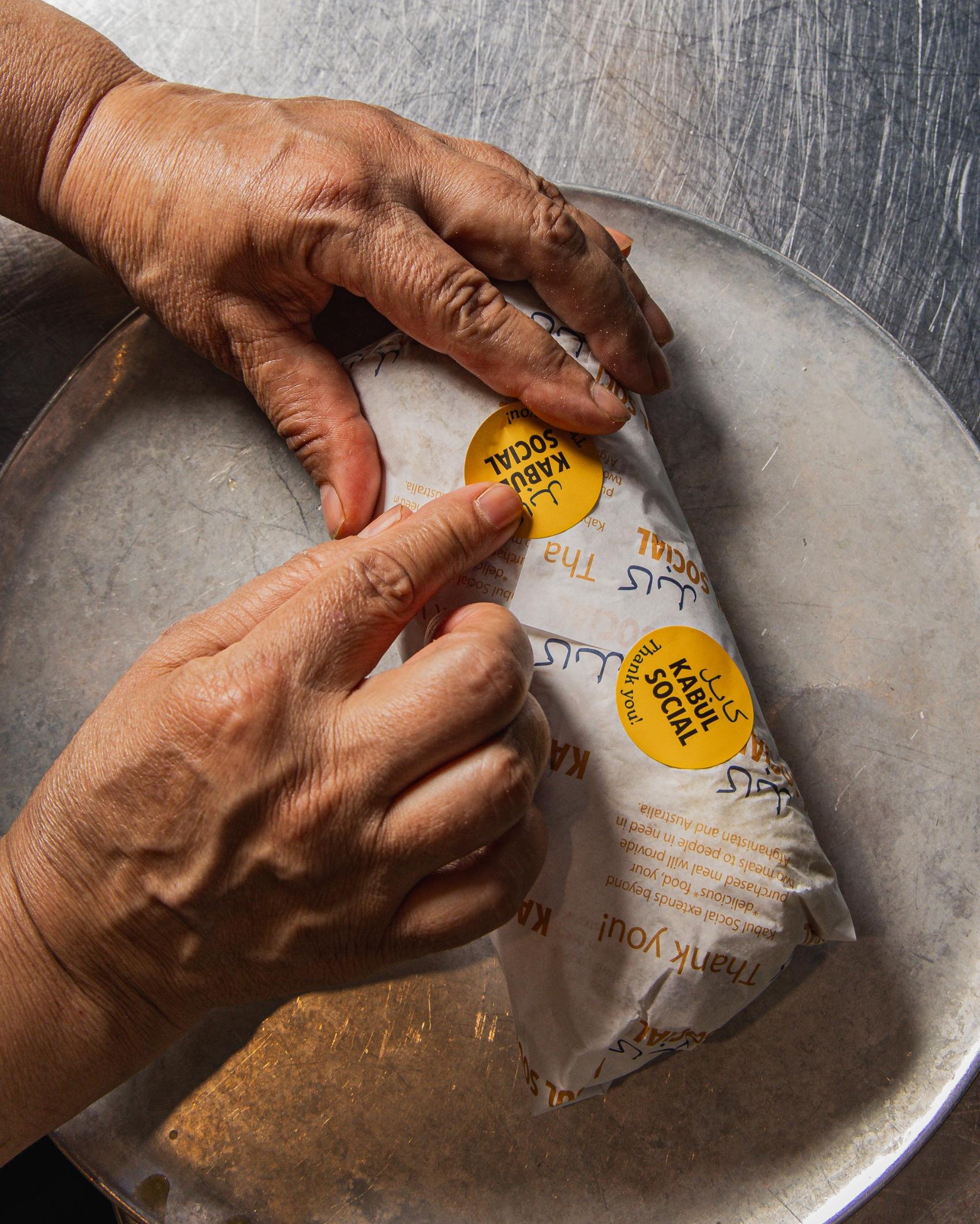 A person wrapping a sandwich in branded paper with yellow and black stickers that read "Kabul Social" and "Thank you" over a metal tray.