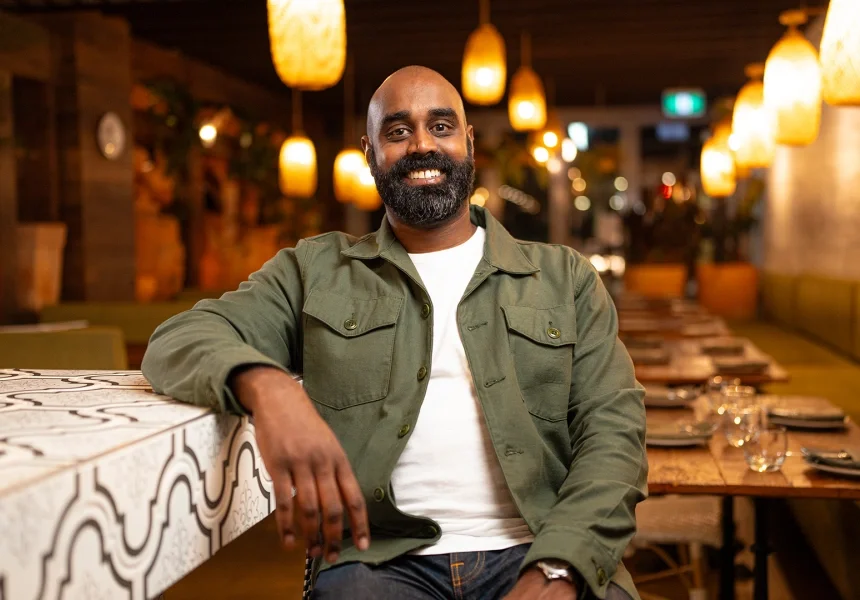 A smiling man with a beard sitting at a table in a cozy restaurant with warm lighting and wooden decor.