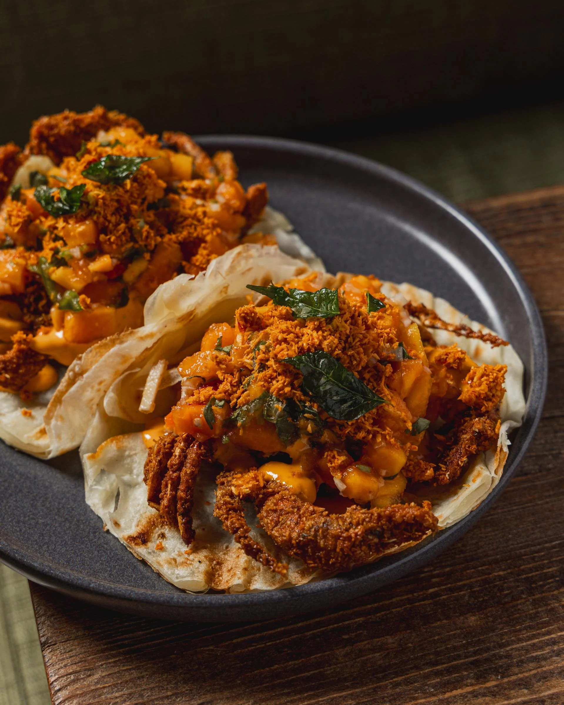 Three crispy fish tacos topped with diced vegetables, shredded cheese, green herbs, and chili flakes, served on a black plate.