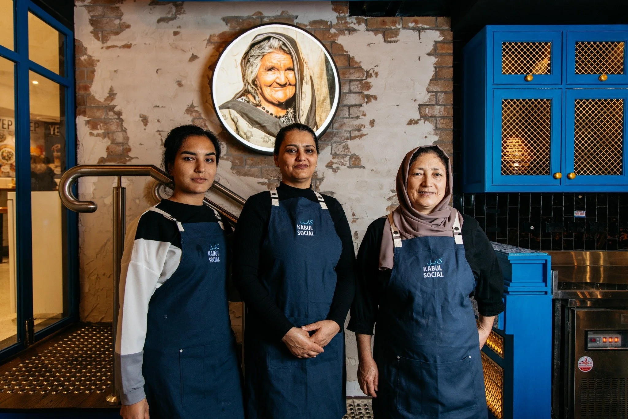 Three women standing side by side in a restaurant, wearing navy blue aprons with white text that says 'Kabul Social', in front of a brick wall with a portrait of an elderly woman displayed above them. The woman on the right is wearing a beige hijab, 