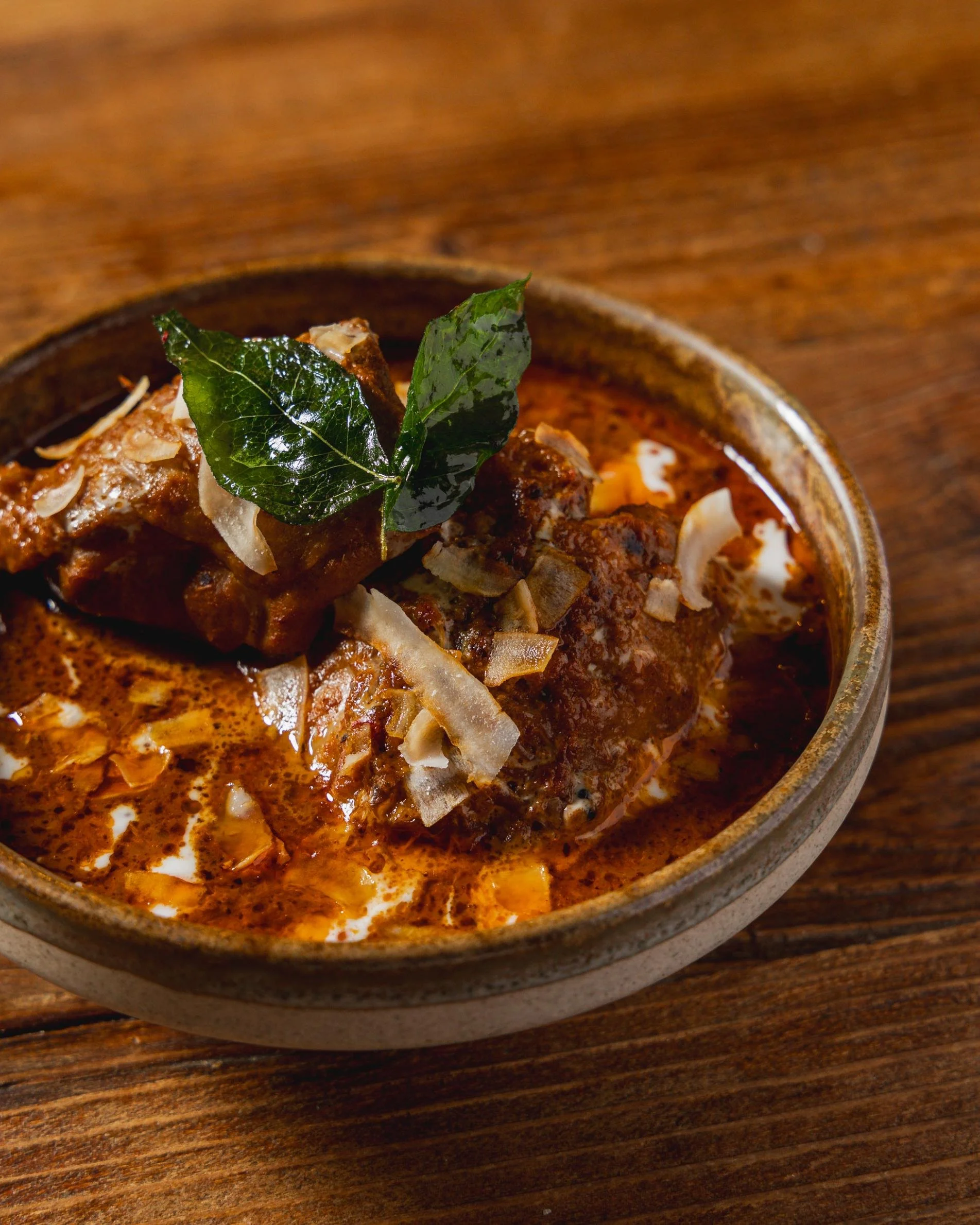 A bowl of Indian curry garnished with a green leaf and shredded coconut, placed on a wooden surface.