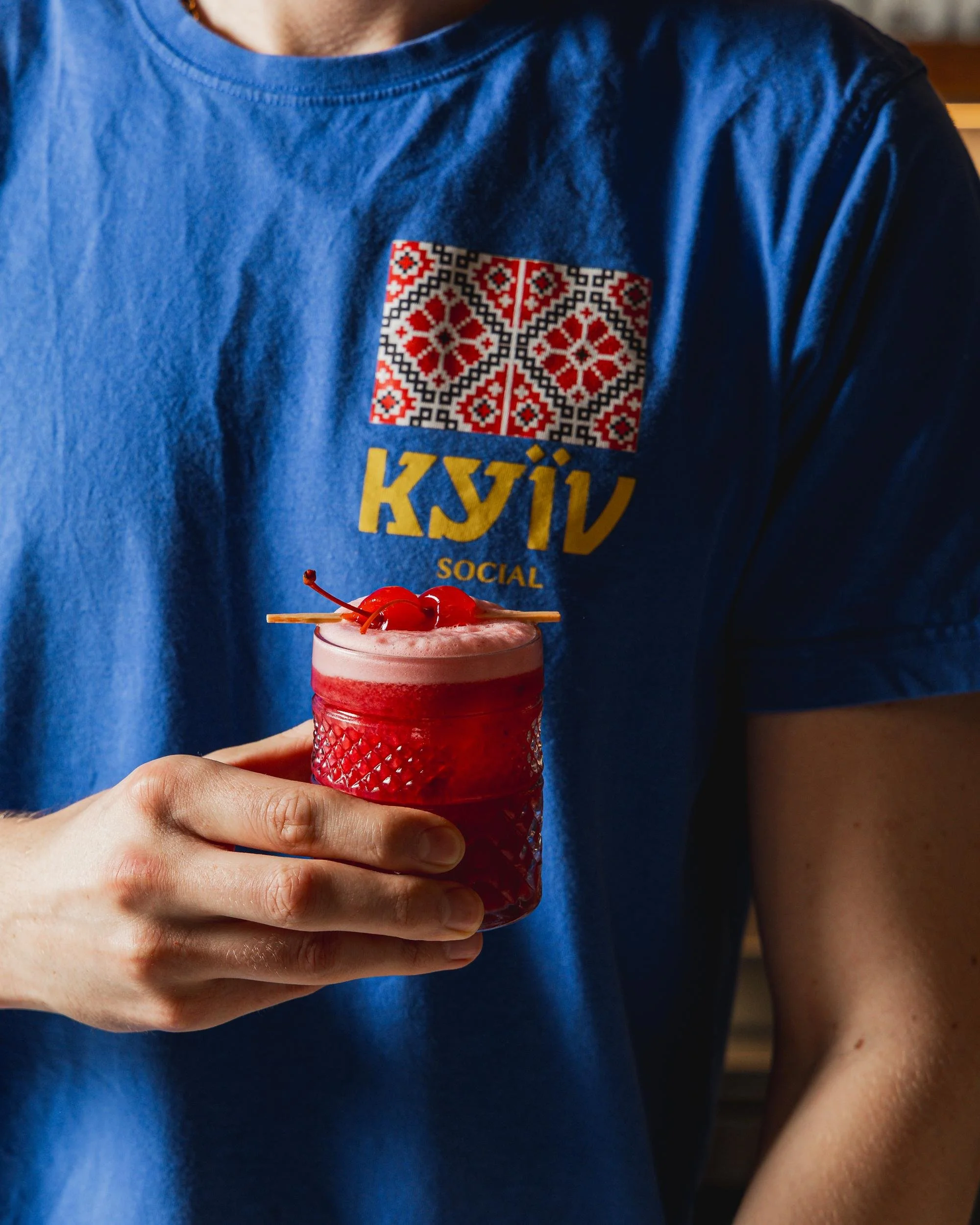 Person wearing a blue shirt with Ukrainian embroidery holding a red cocktail topped with maraschino cherries.