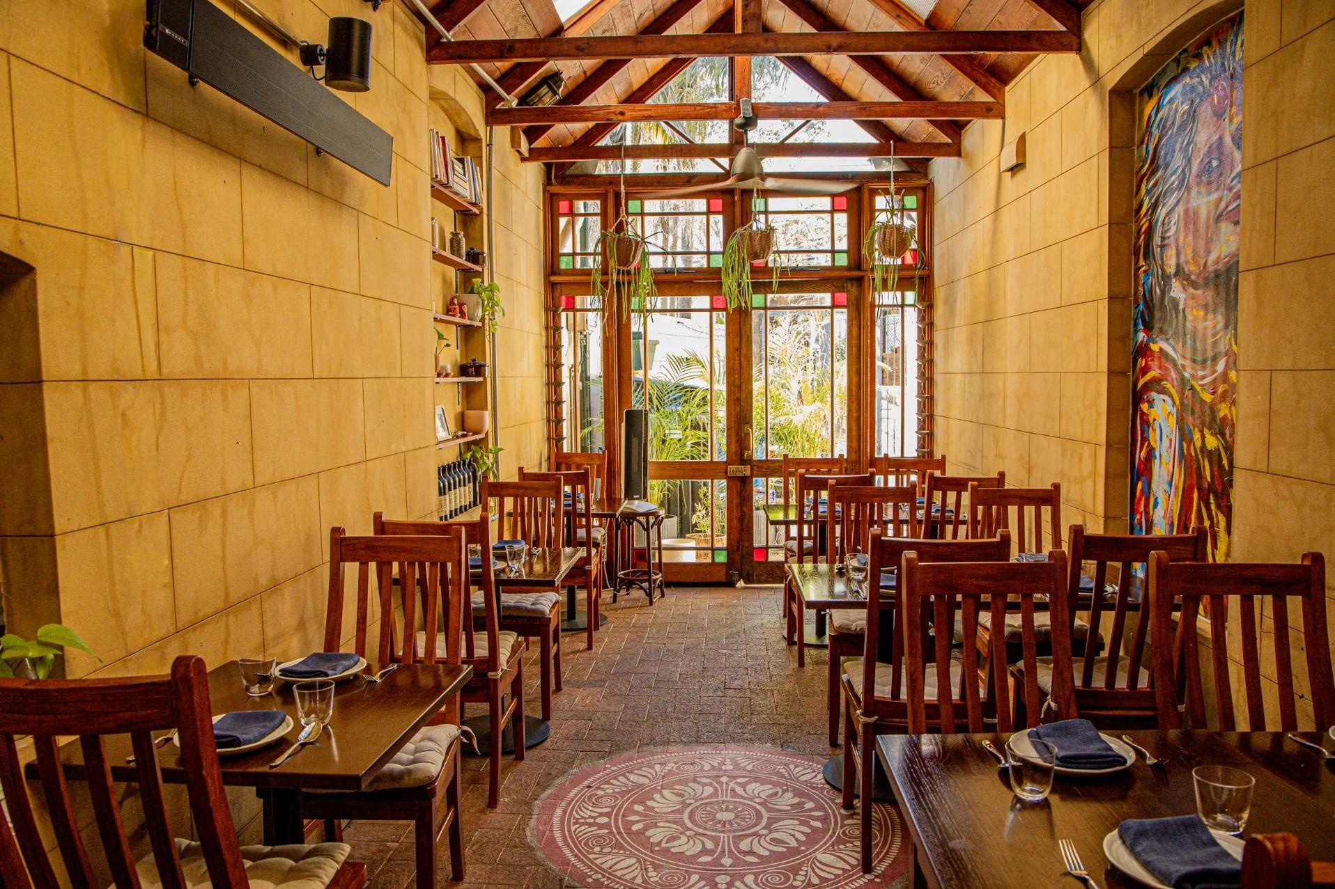 Interior of a warmly lit restaurant with wooden chairs and tables set with blue napkins and glasses, a large colorful abstract painting on the wall, and plants hanging from the ceiling near the front door with stained glass panels.