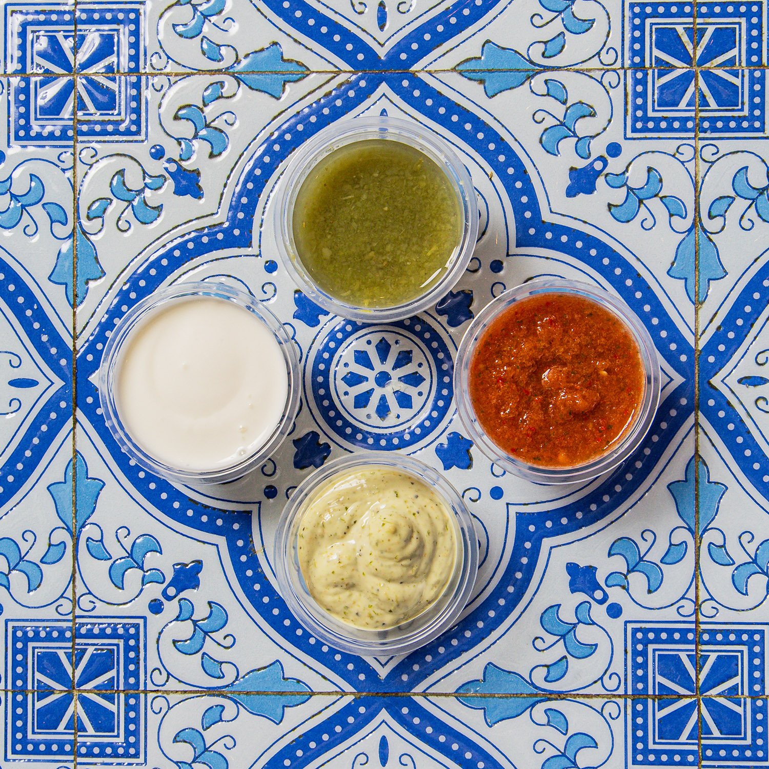 Four small containers of various sauces and condiments placed on a decorative blue and white tiled surface.