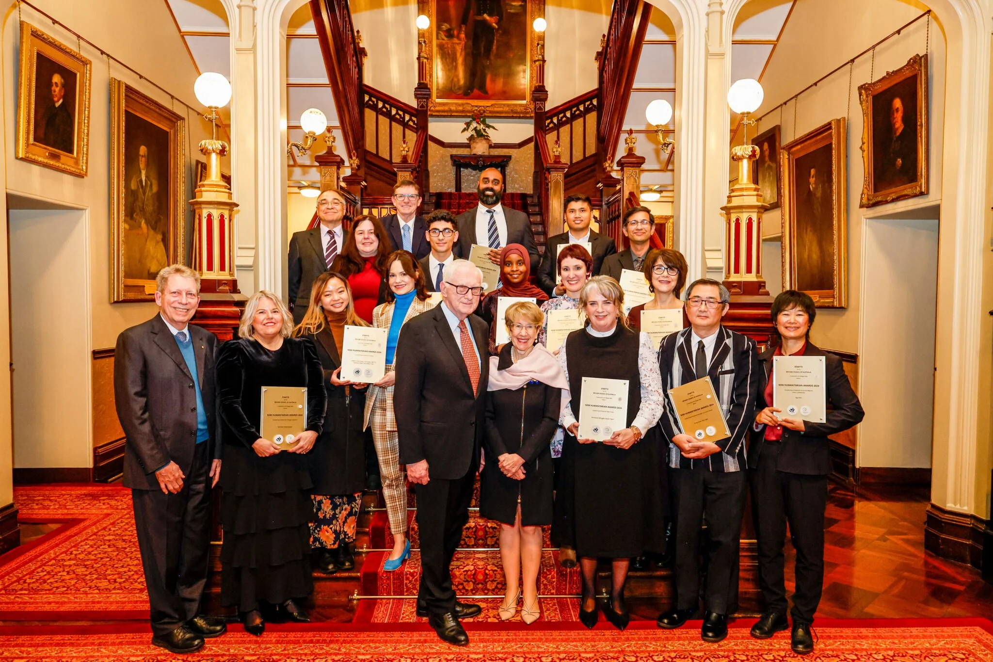 A diverse group of people posing together on a staircase in an elegant, historic building, holding awards and certificates.