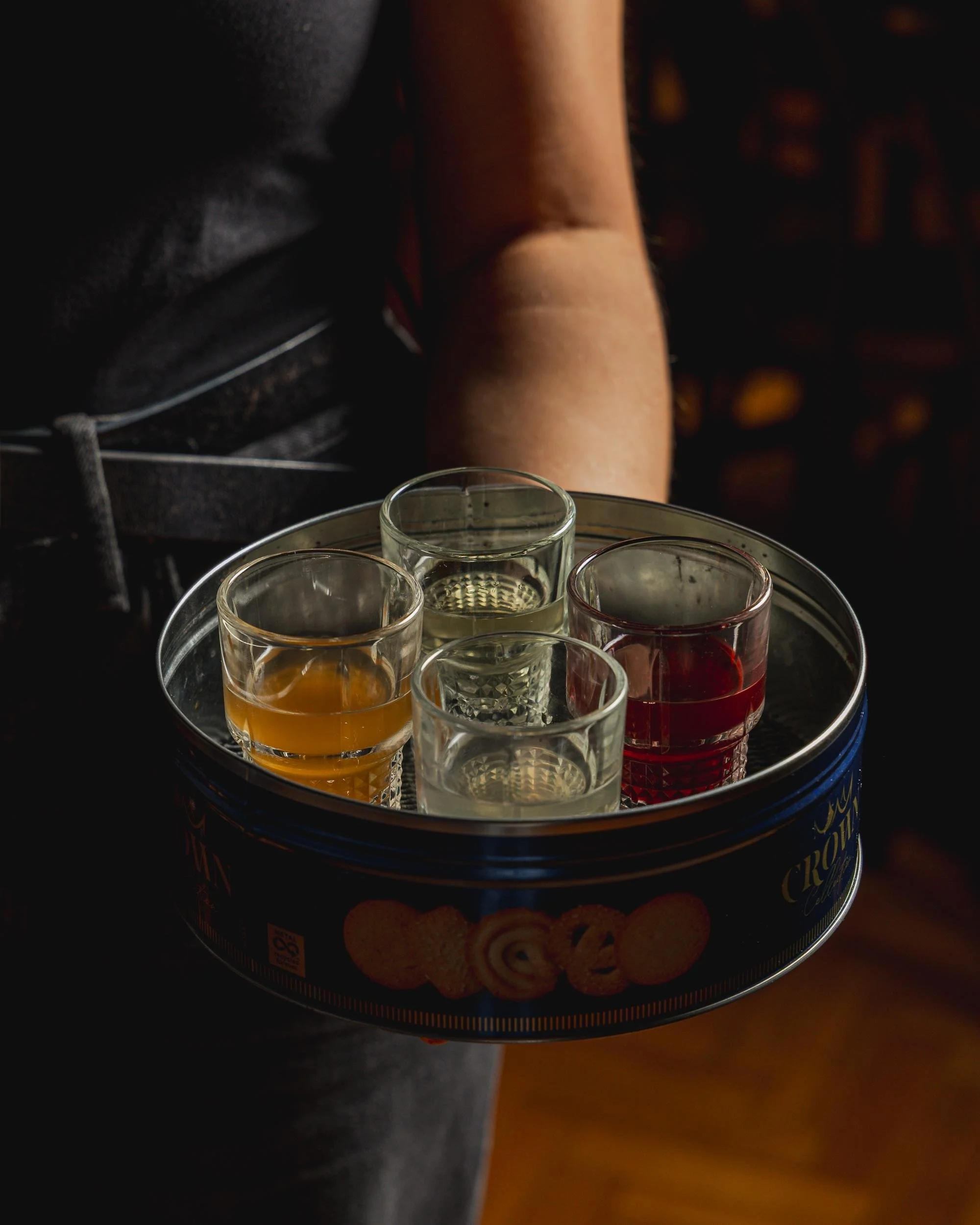 A person holding a round tin tray with four small glasses filled with different colored liquids, possibly alcoholic beverages, inside a dimly lit room.
