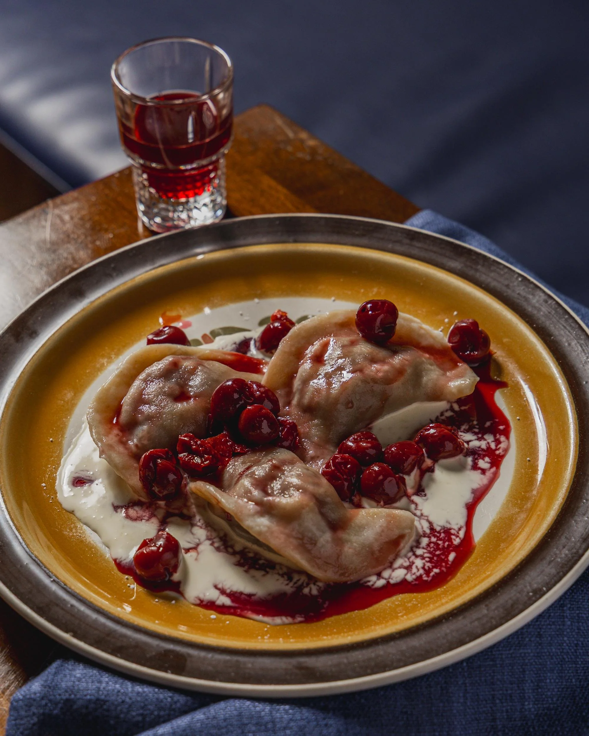 A plate of three pierogis topped with cherries and a drizzle of cherry sauce, served with sour cream, with a glass of red drink on a wooden table.