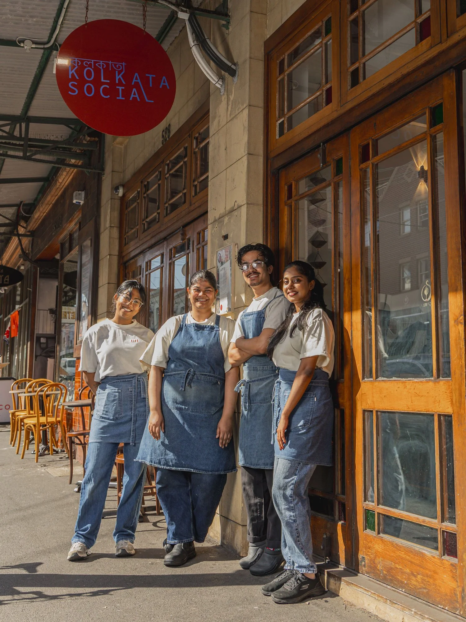 Four staff members standing outside a restaurant with wooden and glass doors, smiling. The restaurant sign reads 'Kolkata Social' in something that looks like Bengali and English.