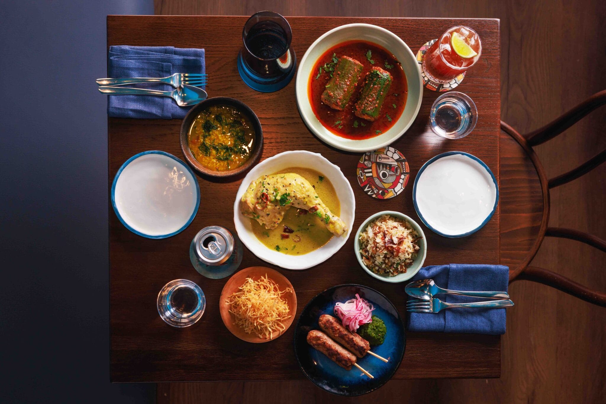 Top-down view of a wooden dining table with various Indian dishes, drinks, and utensils, including samosas, curry, rice, and beverages.