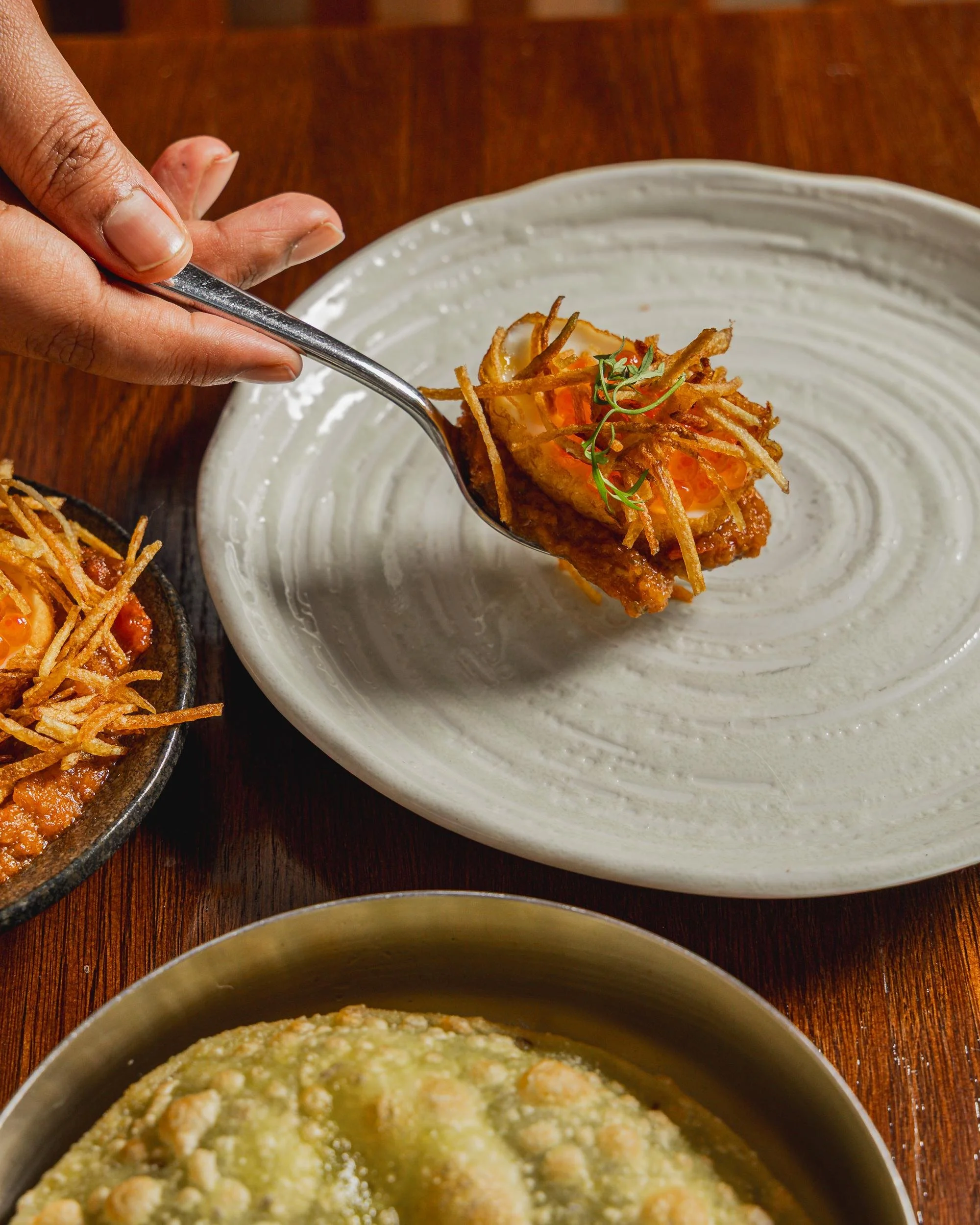 Close-up of a hand holding a spoon with a piece of fried food topped with fried noodles and garnished with microgreens, on a white plate. There are other dishes on a wooden table.