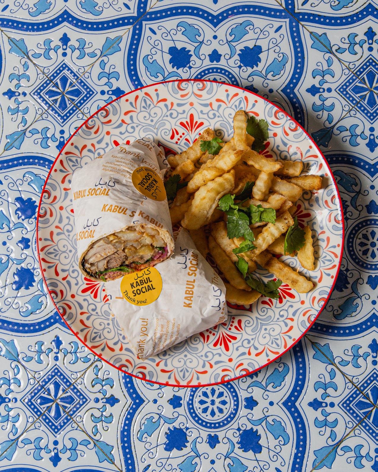 A plate with a wrapped wrap sandwich and a serving of fries garnished with cilantro, placed on a blue and white patterned table.