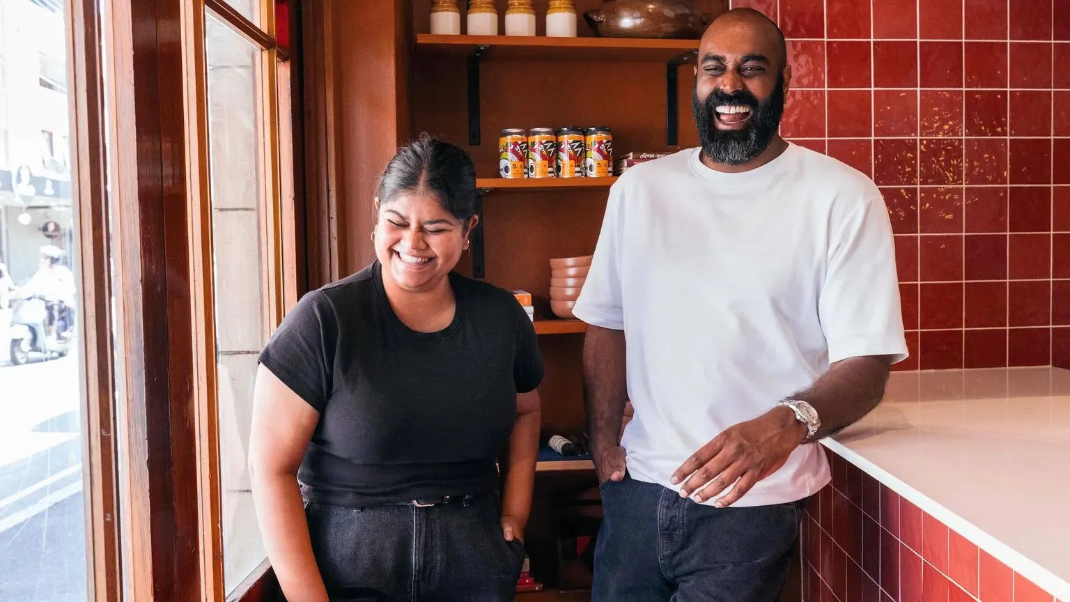 Two people smiling and laughing inside a restaurant, standing near a counter with red tile wall and wooden shelves in the background.