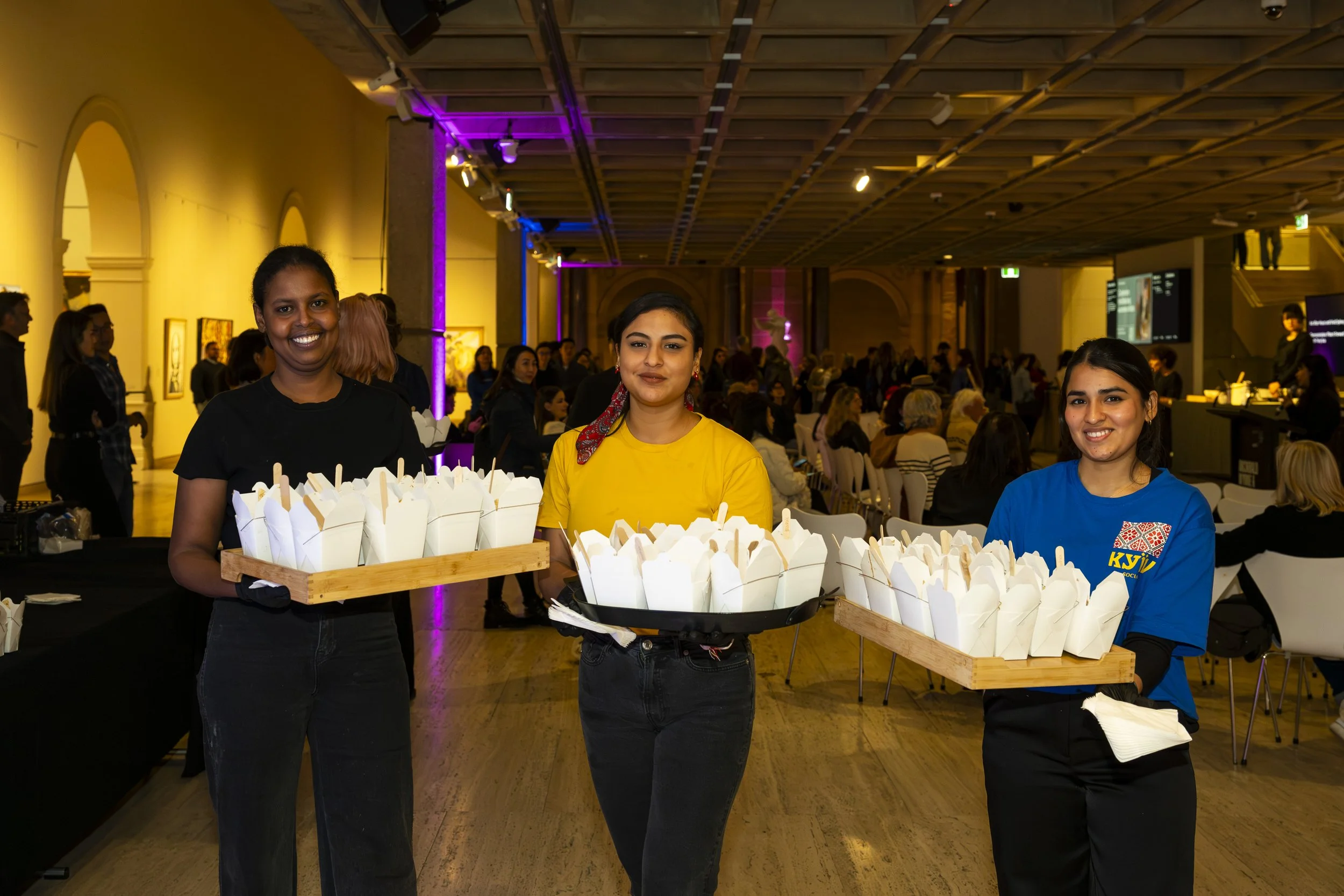 Three women holding trays of food containers in a large event hall with many people seated in the background.