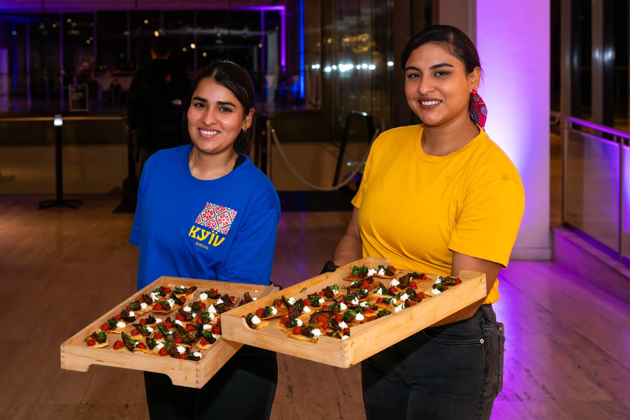 Two women smiling and holding wooden trays with colorful topped flatbreads or appetizers at an indoor event with purple lighting and glass windows in the background.