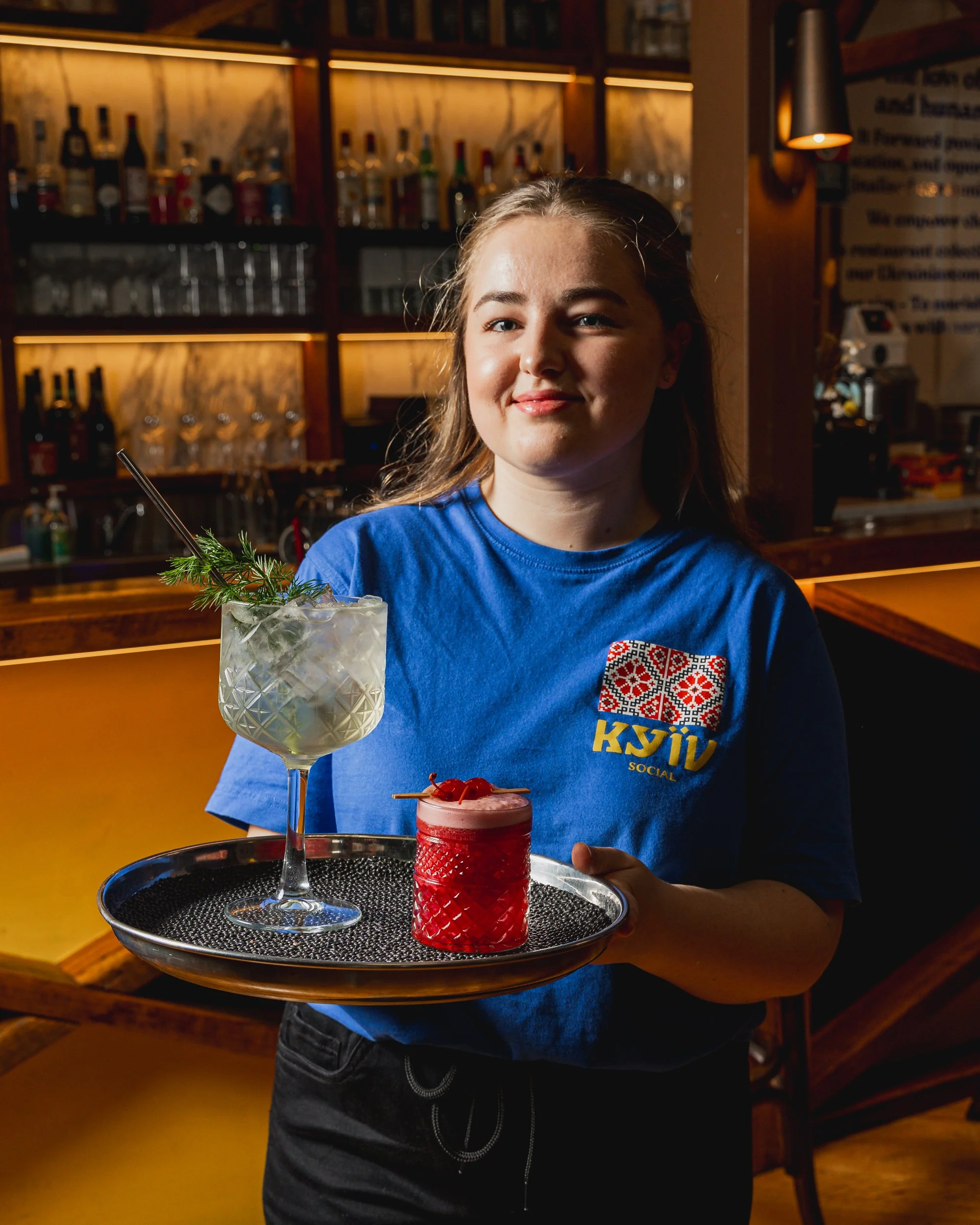 A young woman holding a tray with two cocktails in a bar or restaurant setting, wearing a blue shirt with a logo, smiling slightly.