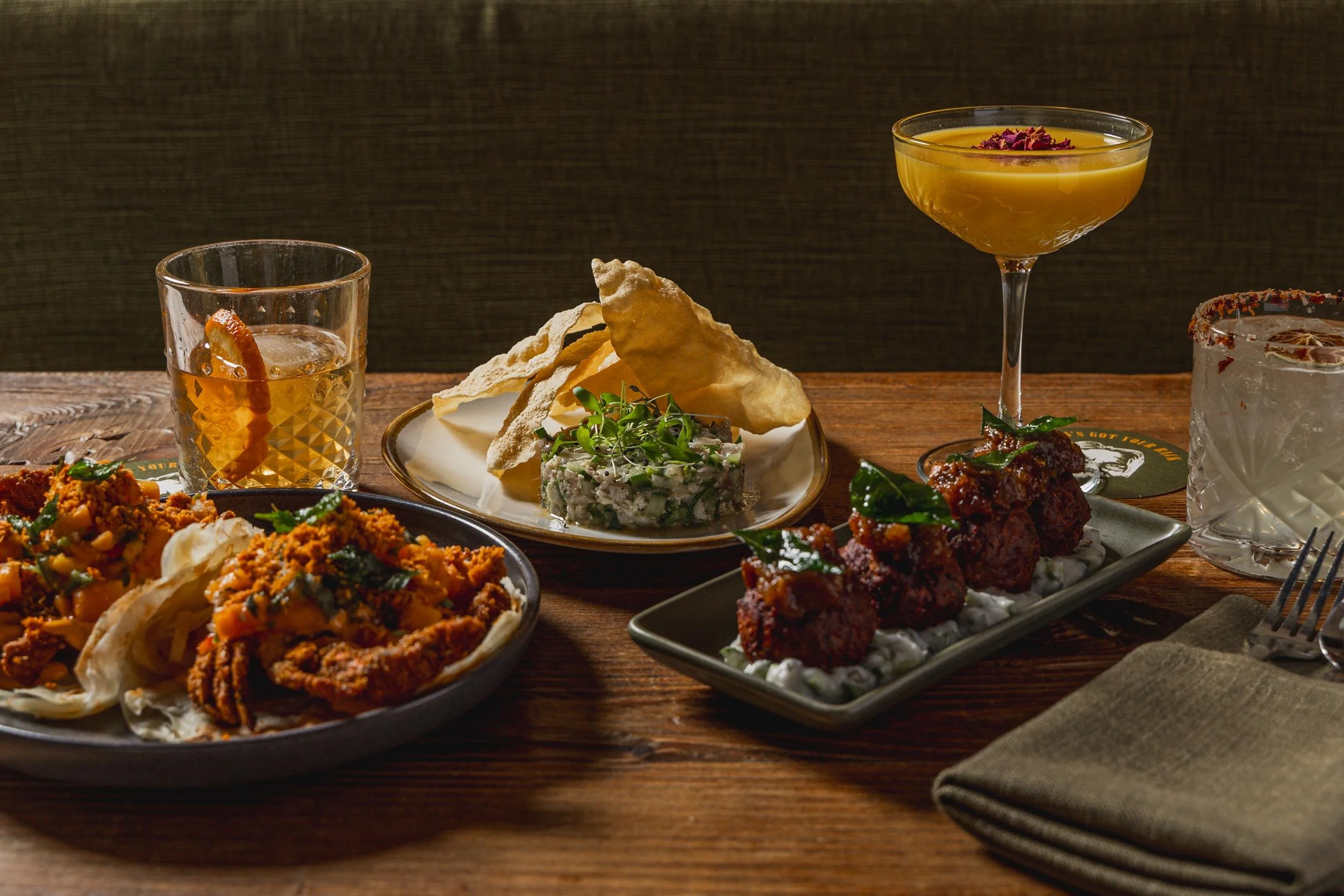 A variety of Indian dishes and drinks on a wooden table, including fried chicken, samosas, chutney, and cocktails.