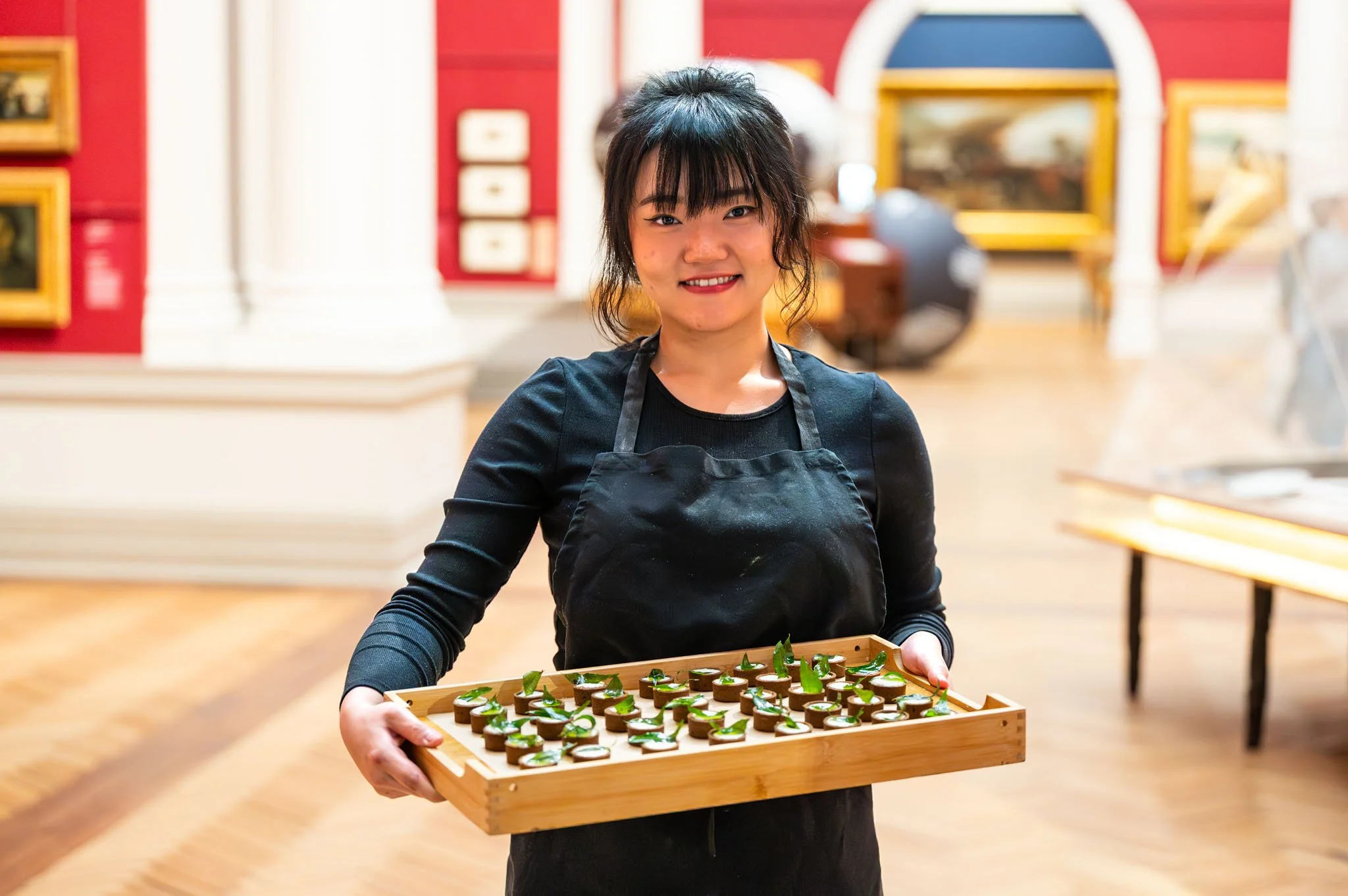 Smiling young woman in black shirt and apron holding a tray of small appetizer bites in a museum.