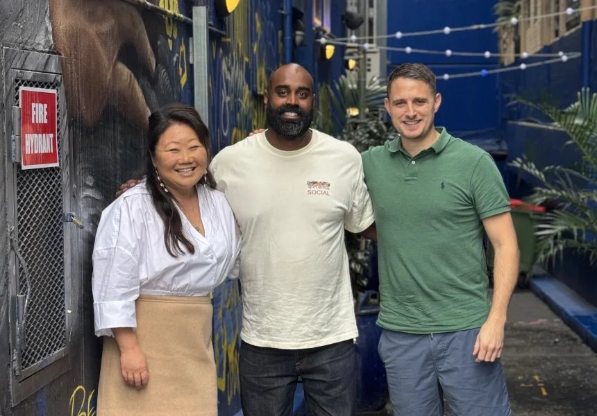 Three people smiling and standing together outdoors in front of a blue wall with graffiti. The person on the left is an Asian woman wearing a white blouse and beige skirt, the person in the middle is a Black man with a beard wearing a white T-shirt, and the person on the right is a Caucasian man with short hair wearing a green polo shirt and blue shorts.