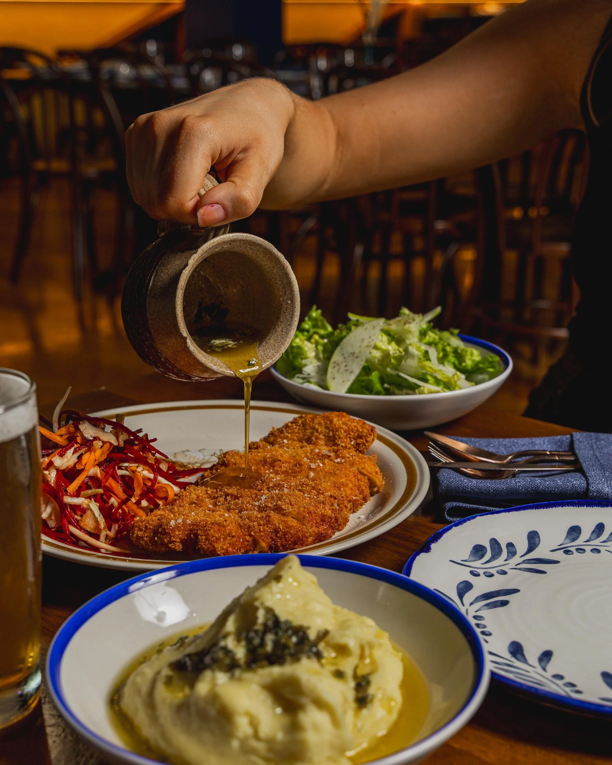 A person pouring gravy over a breaded fried fish fillet with a side of coleslaw and mashed potatoes on a dining table.