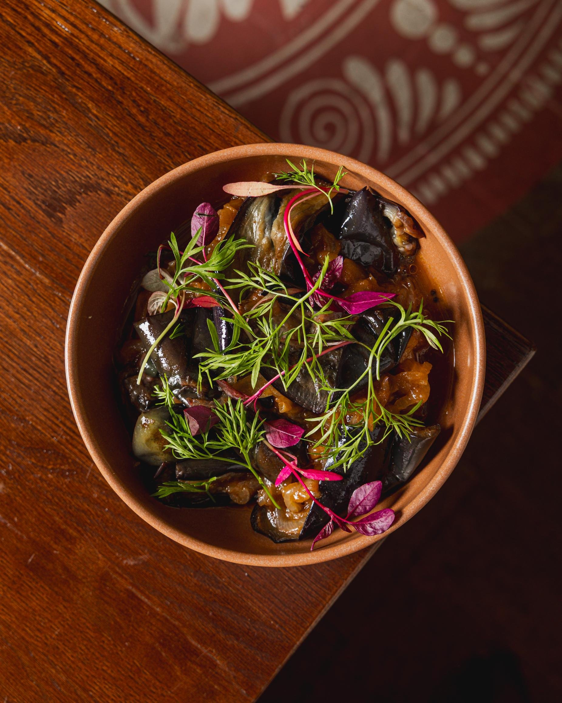 A bowl of eggplant dish garnished with fresh herbs and microgreens, placed on a wooden surface.
