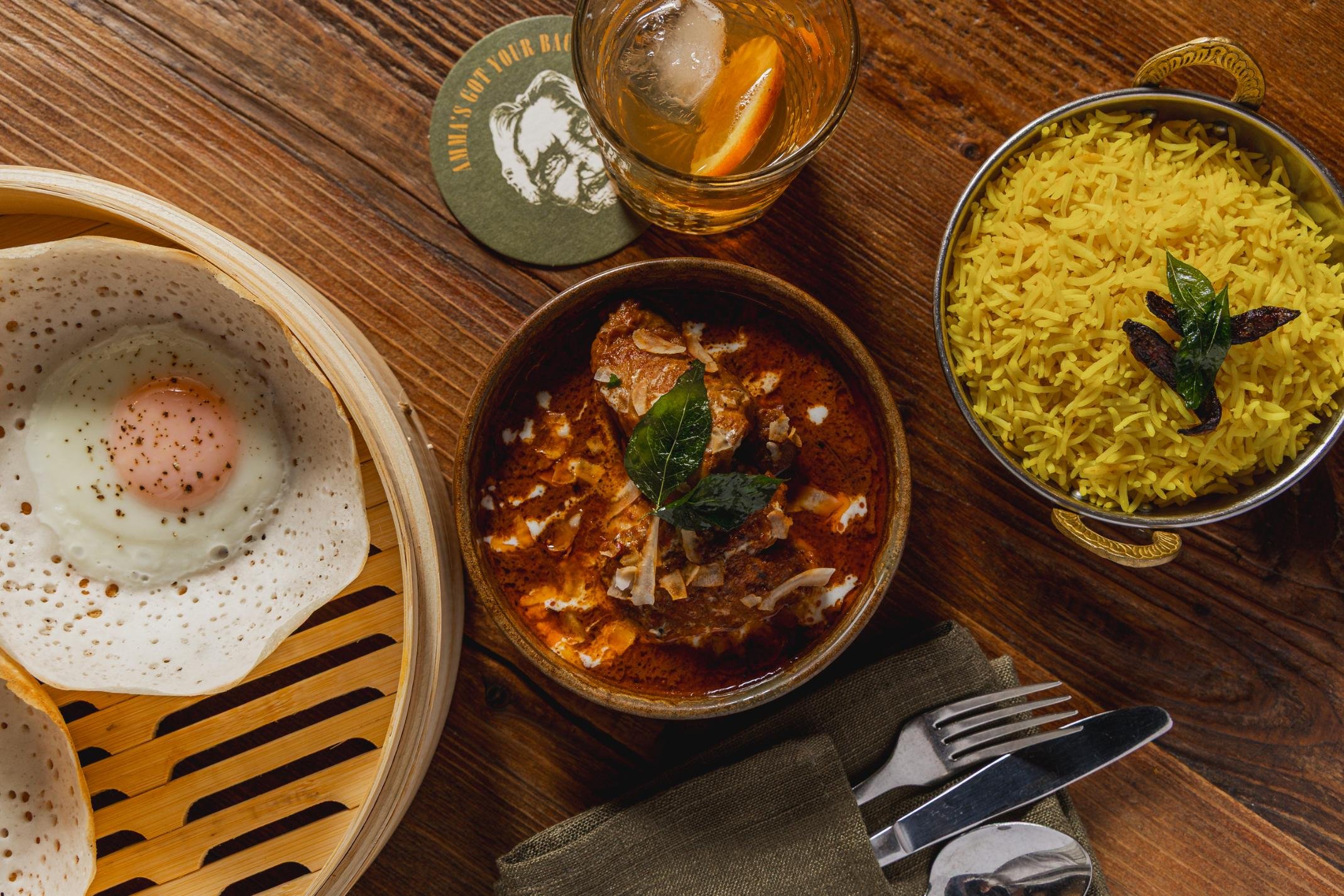 Indian meal with a fried egg, a bowl of chicken curry garnished with bay leaves, a bowl of yellow rice with bay leaves, and a glass of drink on a wooden table.
