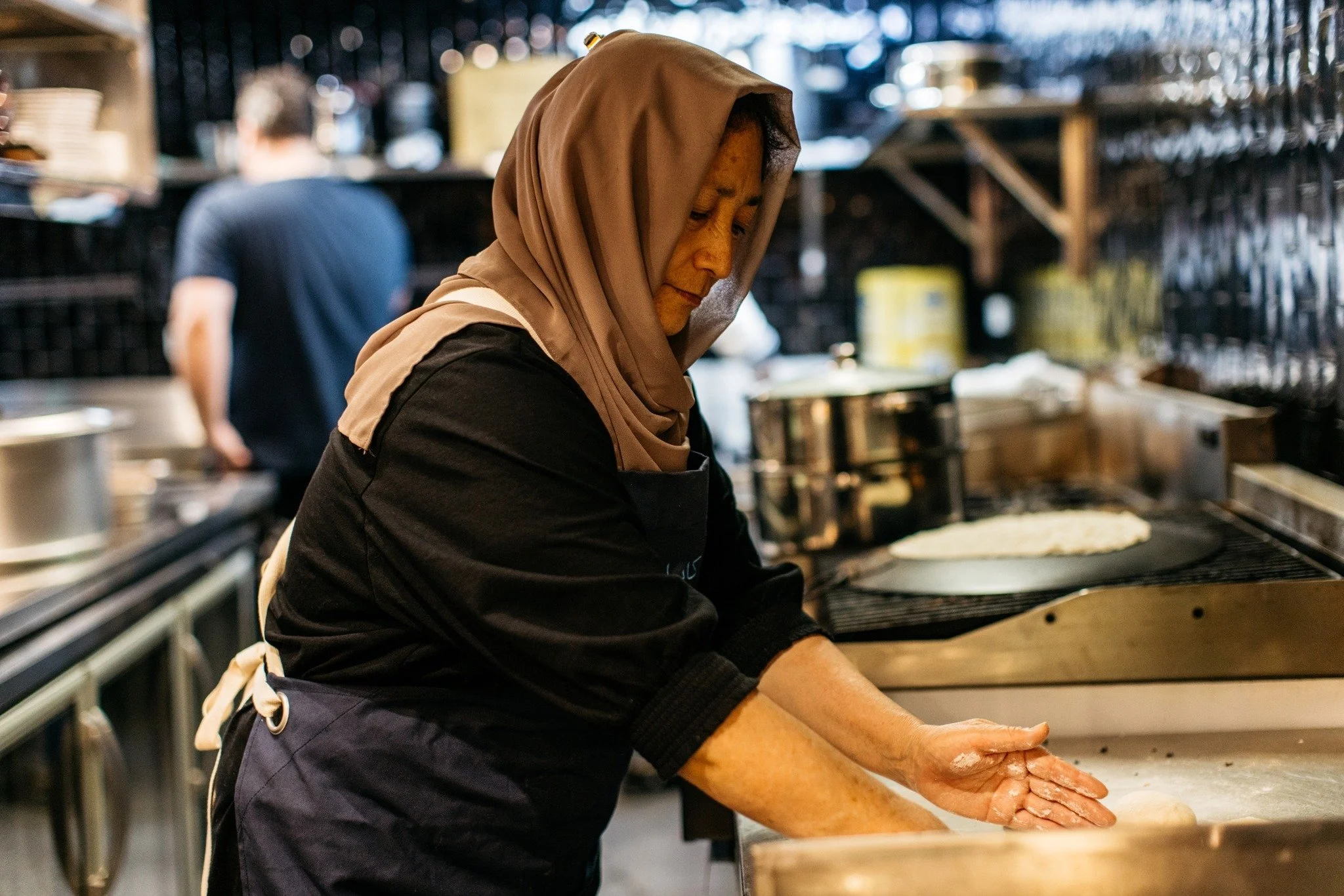 A woman wearing a brown headscarf and black shirt baking bread in a bakery kitchen with a man in the background.