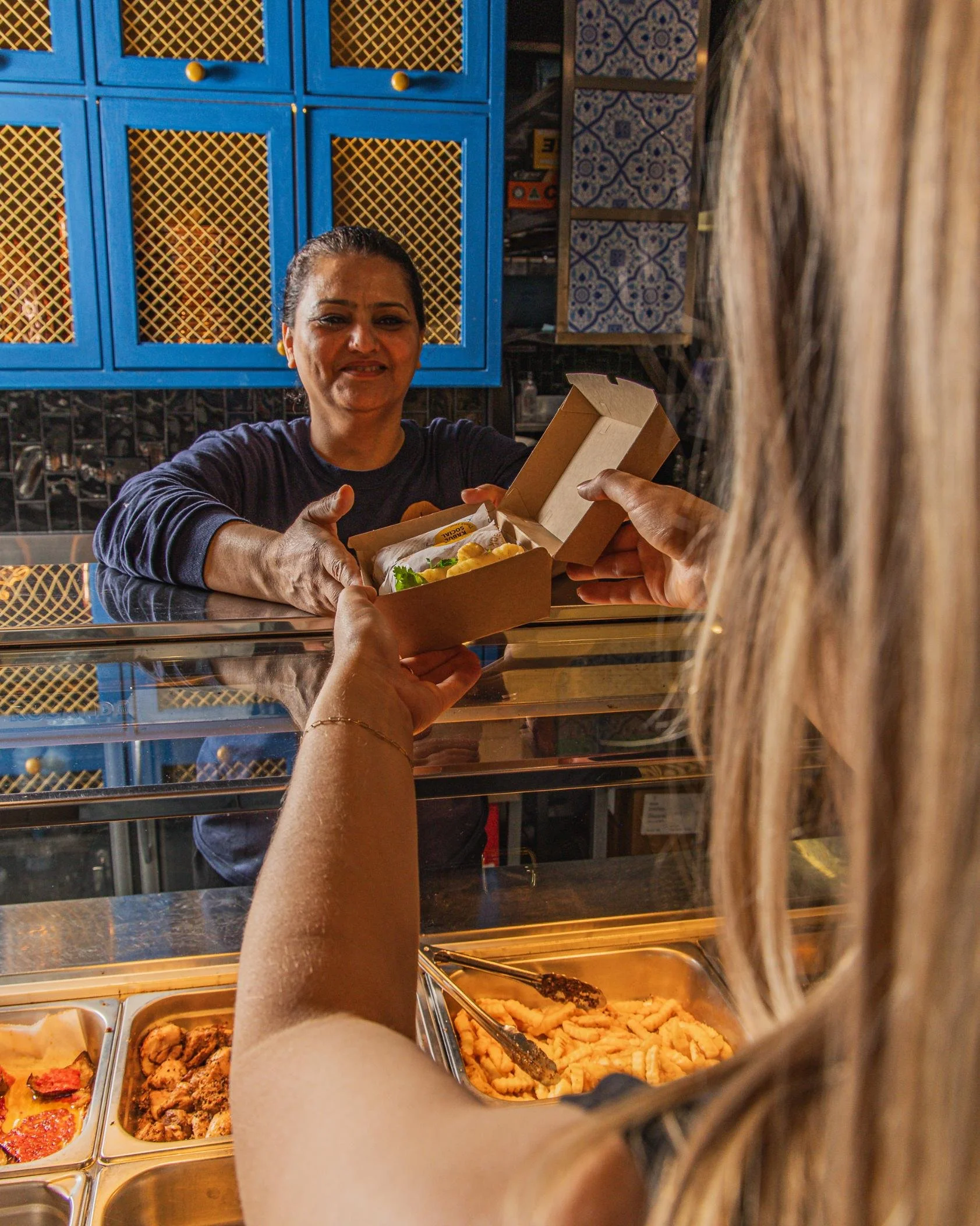 A woman at a food counter handing a box of food to a customer with long blonde hair.