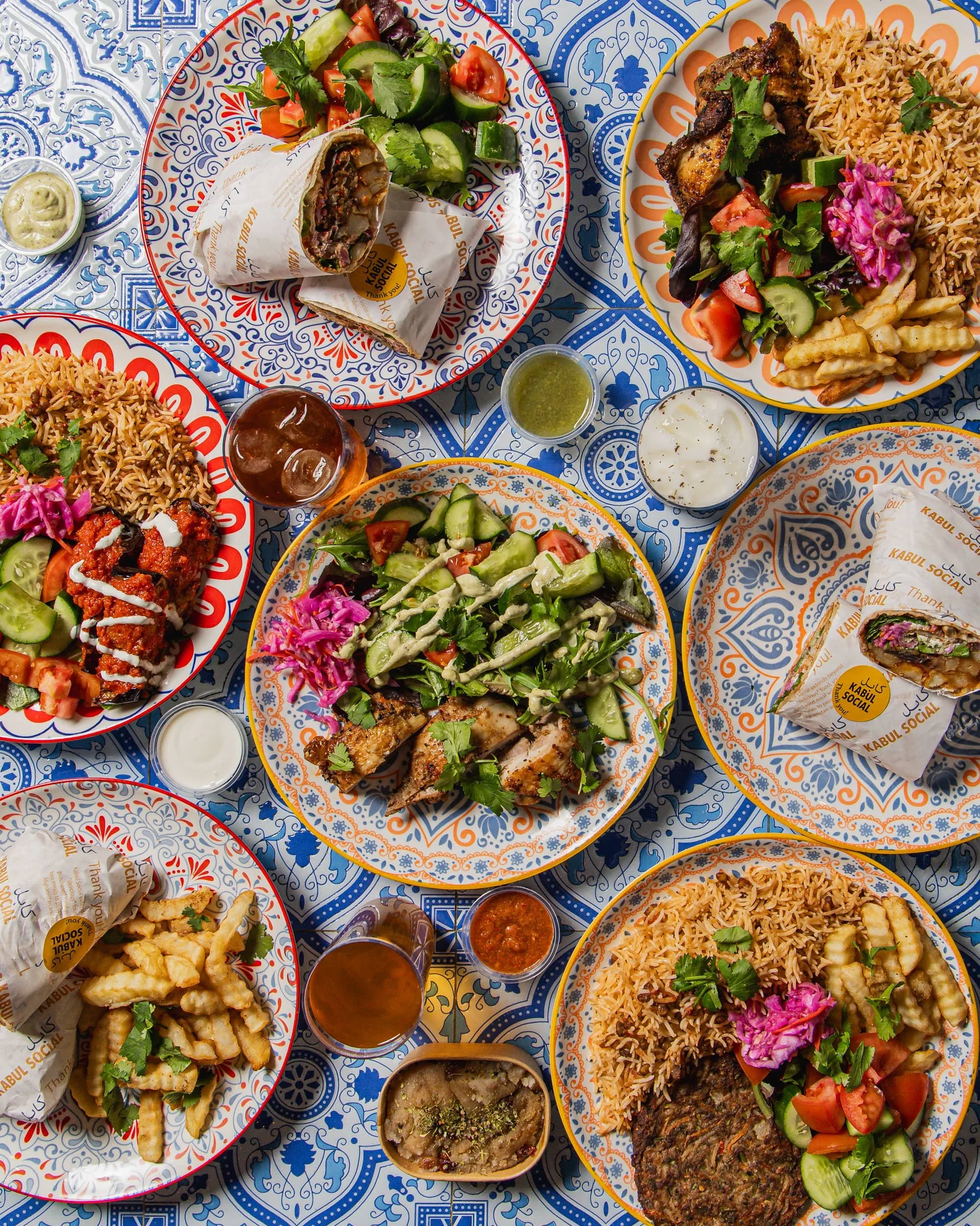 A colorful table with various Middle Eastern dishes including salads, rice, grilled meats, wraps, fries, sauces, and a bowl of soup, set on decorative plates and small bowls.