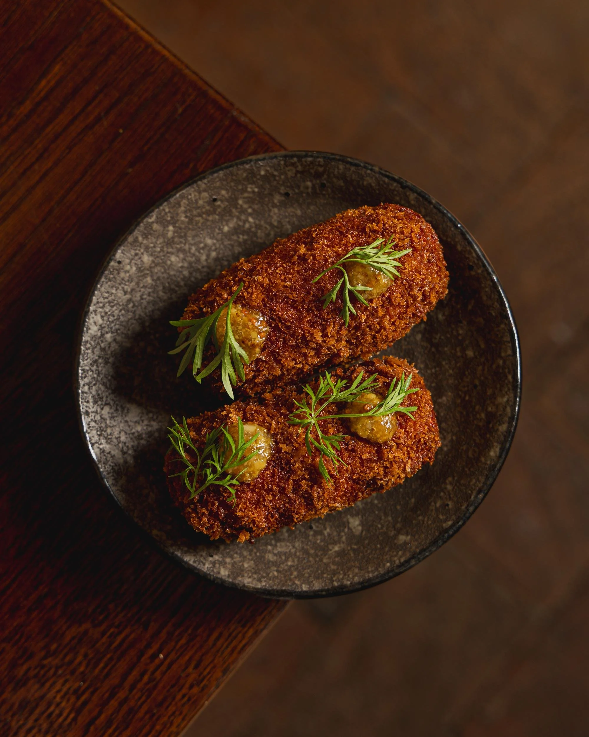 Two breaded and fried croquettes garnished with green herbs and mustard on a dark ceramic plate.