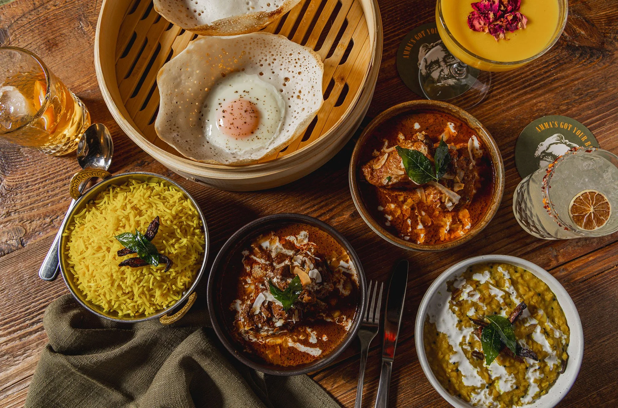 A variety of Indian dishes on a wooden table, including bowls of curry, rice, and a fried egg in a bamboo steamer, along with drinks like lemon water and a cocktail garnished with rose petals.