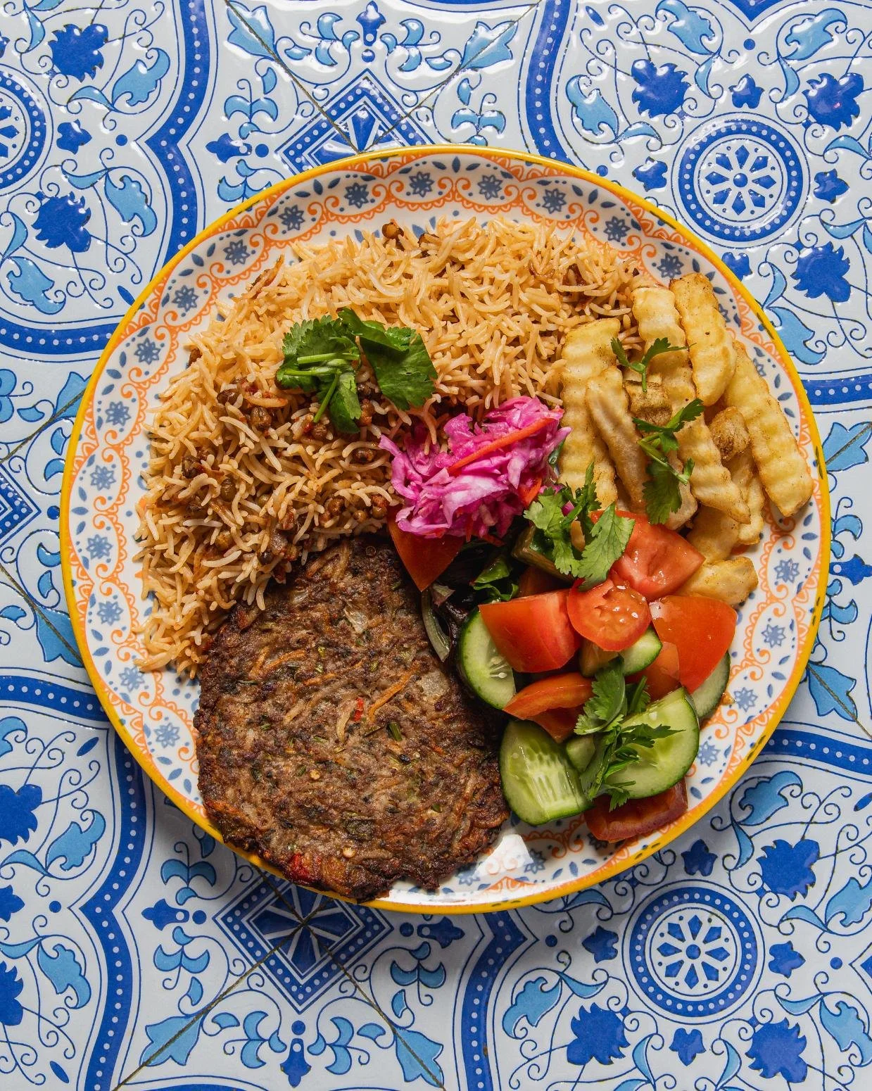 Plate with rice, fried fish fillet, salad with cucumbers, tomatoes, cilantro, pickled cabbage, and fried plantains, on a blue and white patterned tablecloth.