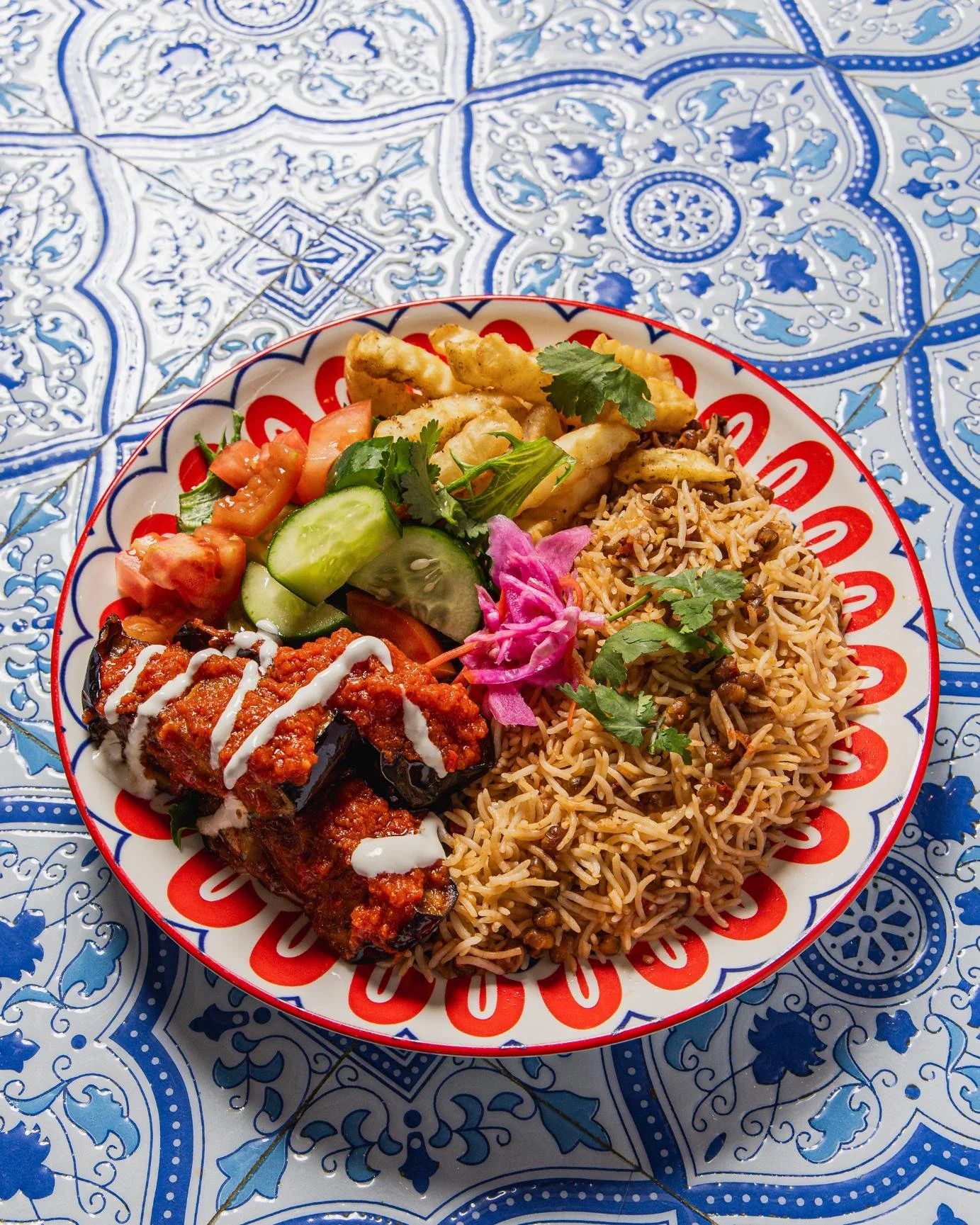 Plate of mixed Indian dishes on a decorative blue and white tiled table, including bhel puri, vegetable salad, eggplant with yogurt sauce, and fried rice garnished with cilantro and pink pickled onions.