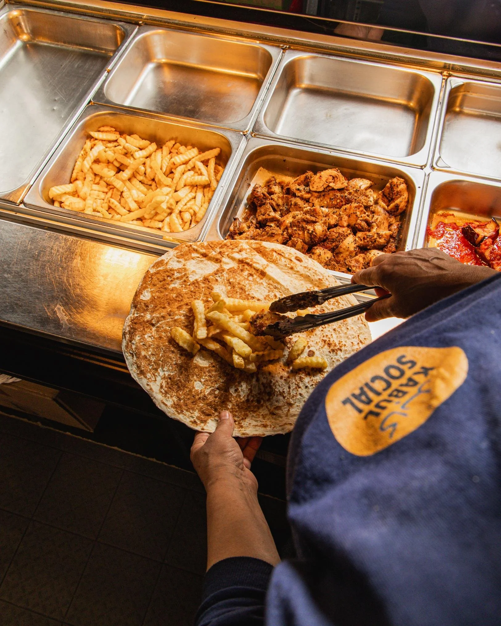 Person serving French fries onto a large toasted bun at a buffet-style food counter with trays of French fries, seasoned chicken, and sauce.