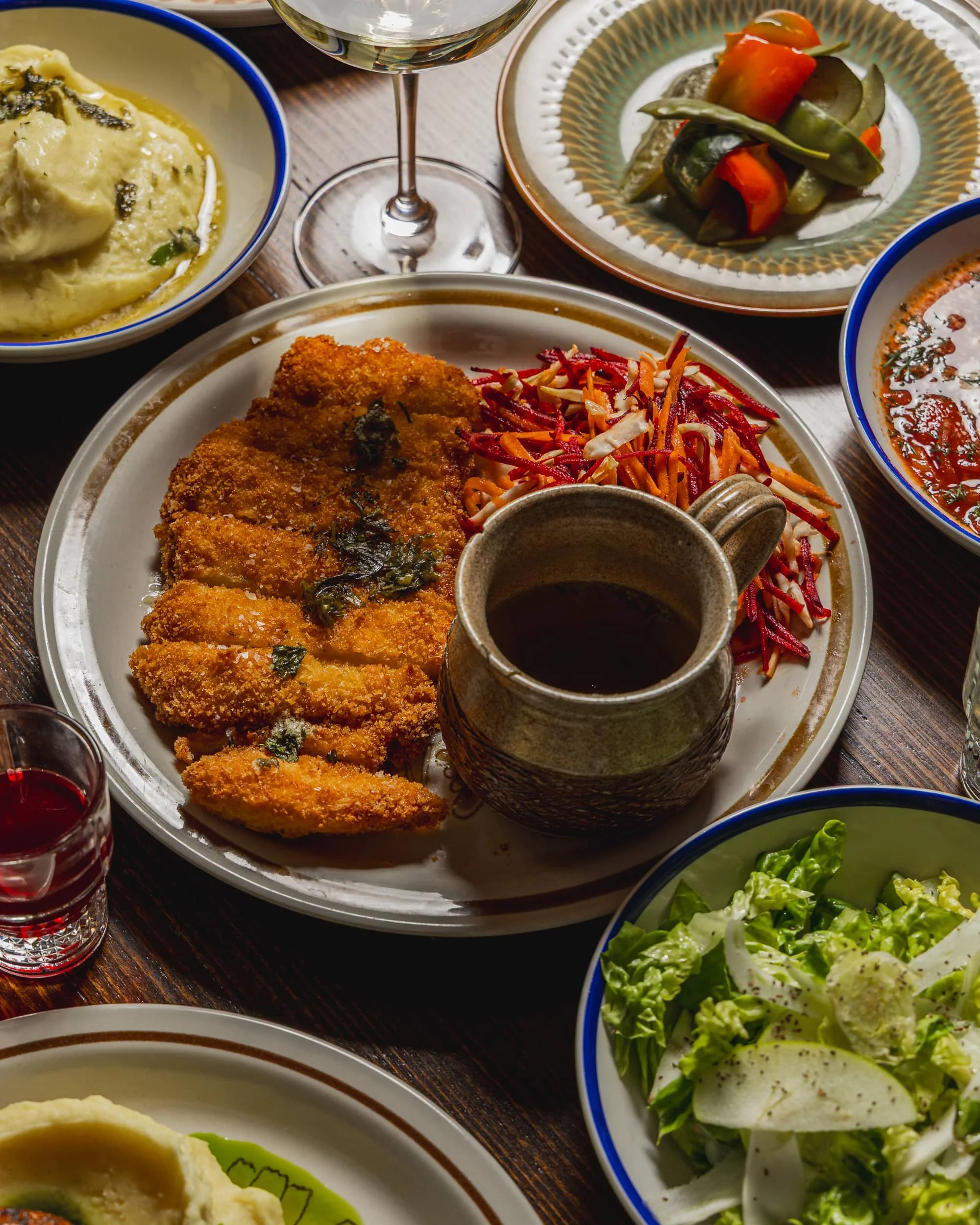 A plate of breaded fried chicken with coleslaw and a small pitcher of black coffee on a table surrounded by various side dishes including salad, mashed potatoes, vegetables, and a glass of white wine.