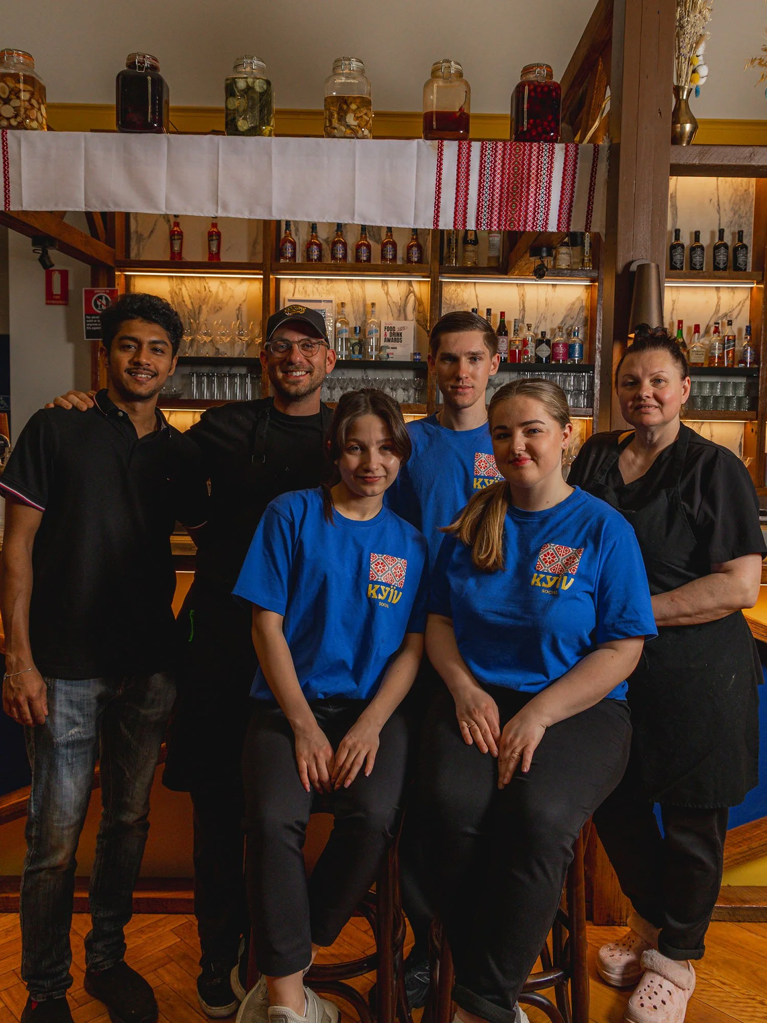 Group of six people, including two women and four men, smiling and posing in a bar or restaurant, with shelves of bottles and jars behind them.