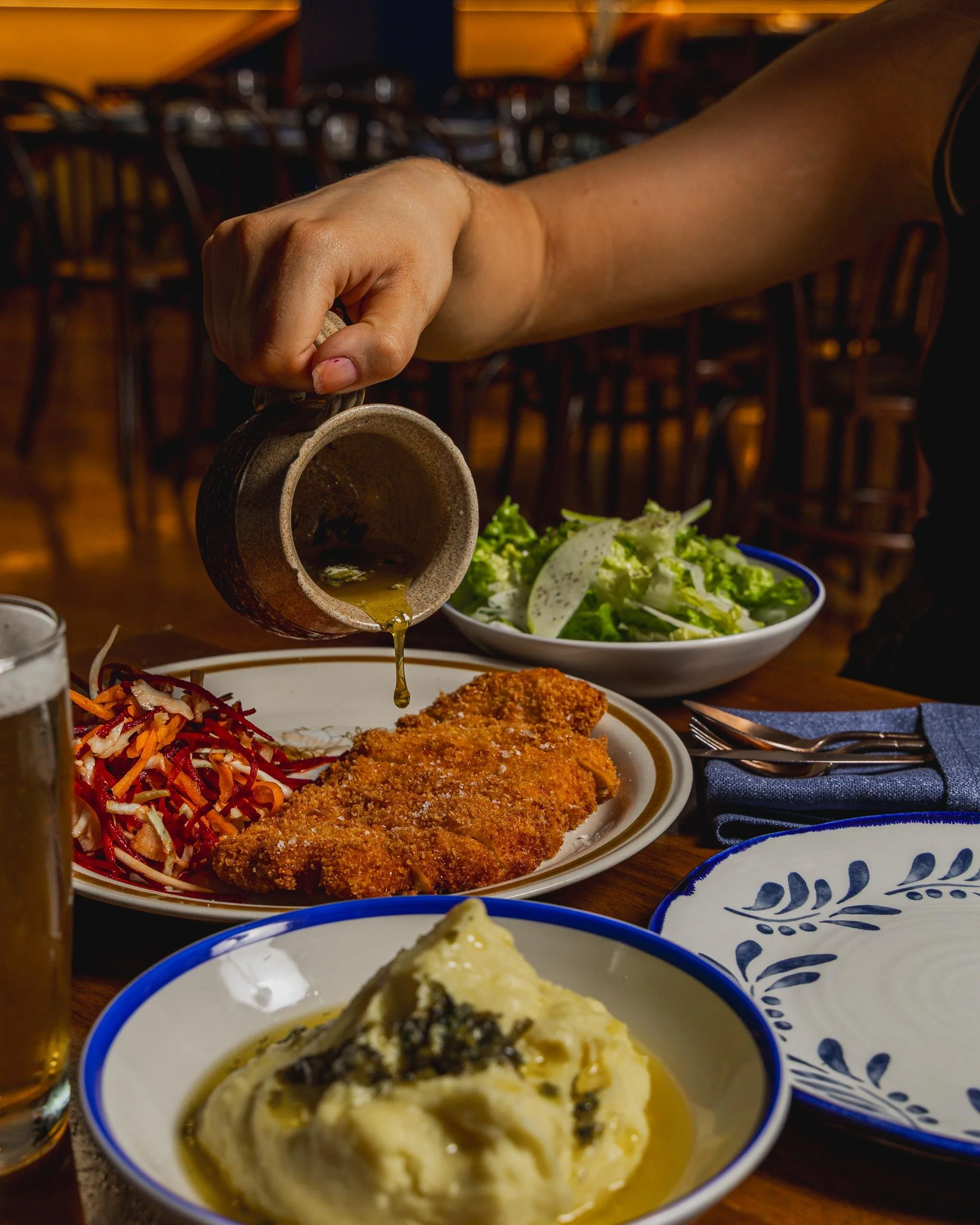 Person pouring gravy over a breaded fried fish fillet with side dishes of coleslaw, mashed potatoes with gravy, and a salad in a bowl on a wooden table.