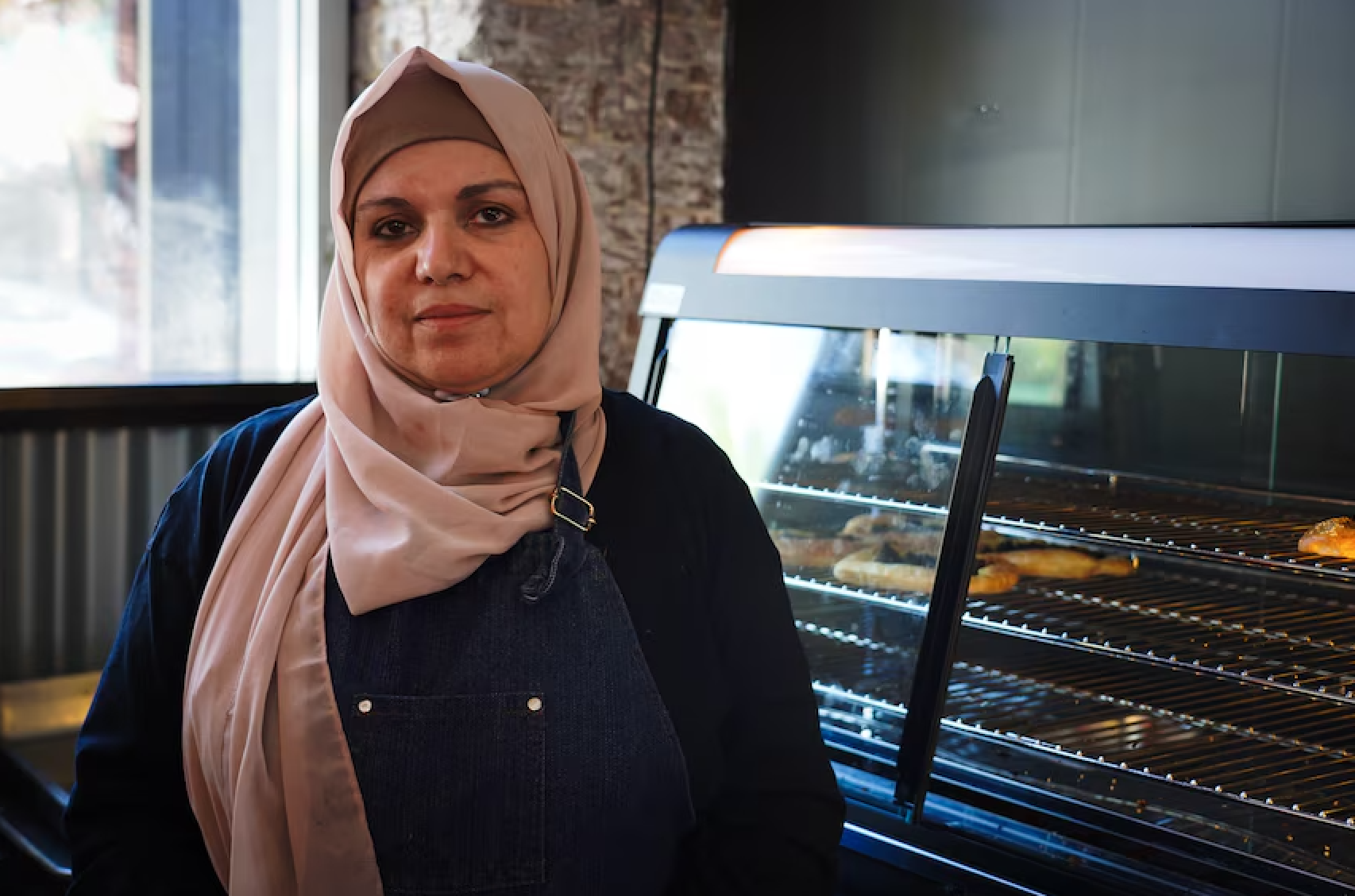 A woman wearing a beige hijab and black apron standing next to a glass display case with baked goods inside.