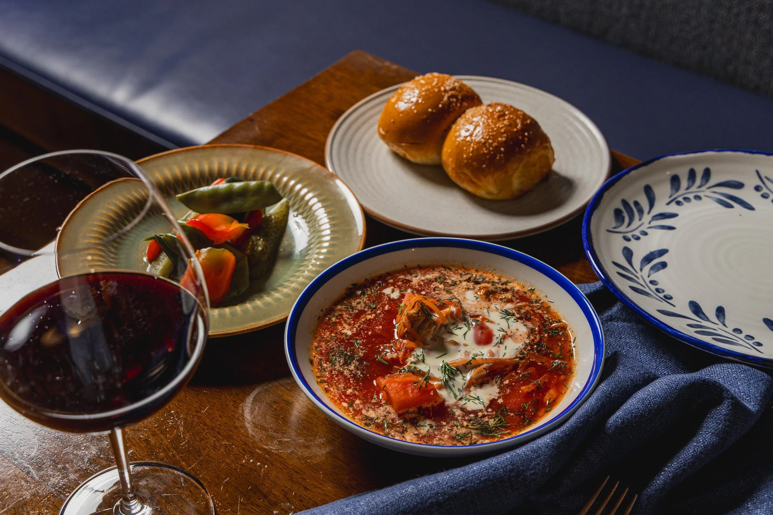 A table set with a bowl of tomato soup topped with herbs and a dollop of cream, a plate of bread rolls, a plate of pickled vegetables, and glasses of red wine.
