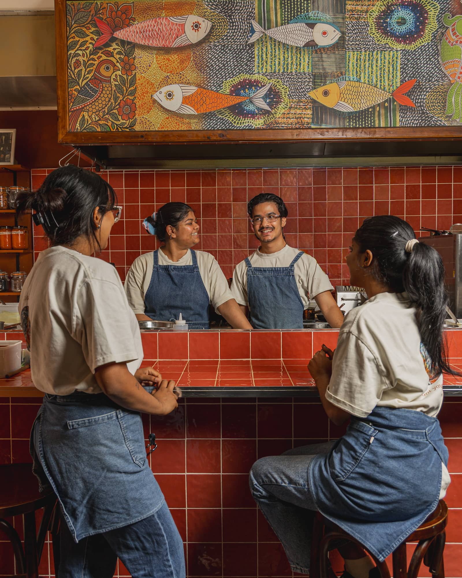 Four people, two women and one man, are standing and sitting at a counter in a restaurant, wearing white t-shirts and denim aprons, engaging in a conversation with two women who are standing. The background features small red tiles and a colorful fis