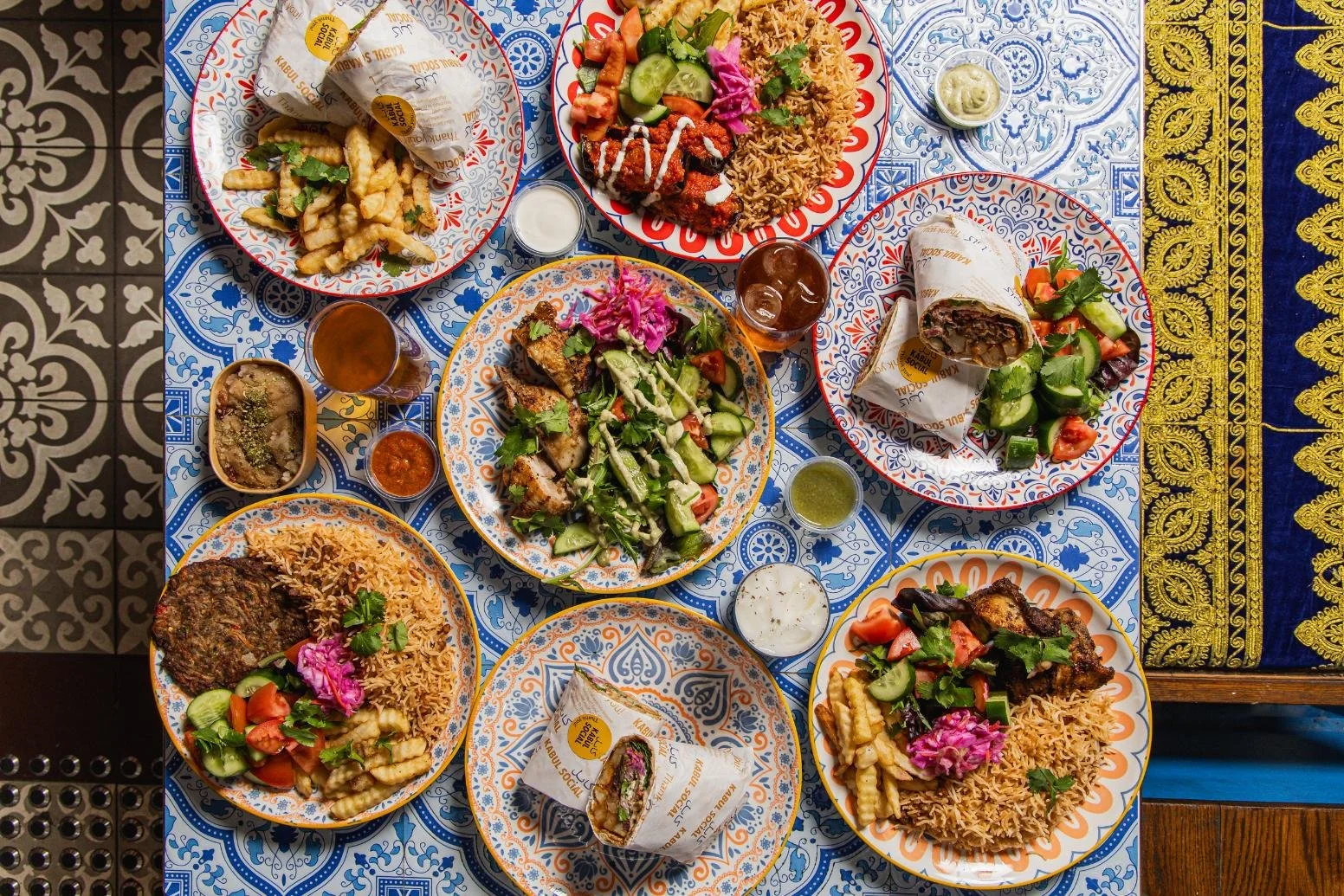 A table with multiple plates of Middle Eastern or Mediterranean food, including salads, rice, kebabs, wraps, and various sauces and drinks, set on a patterned tablecloth.