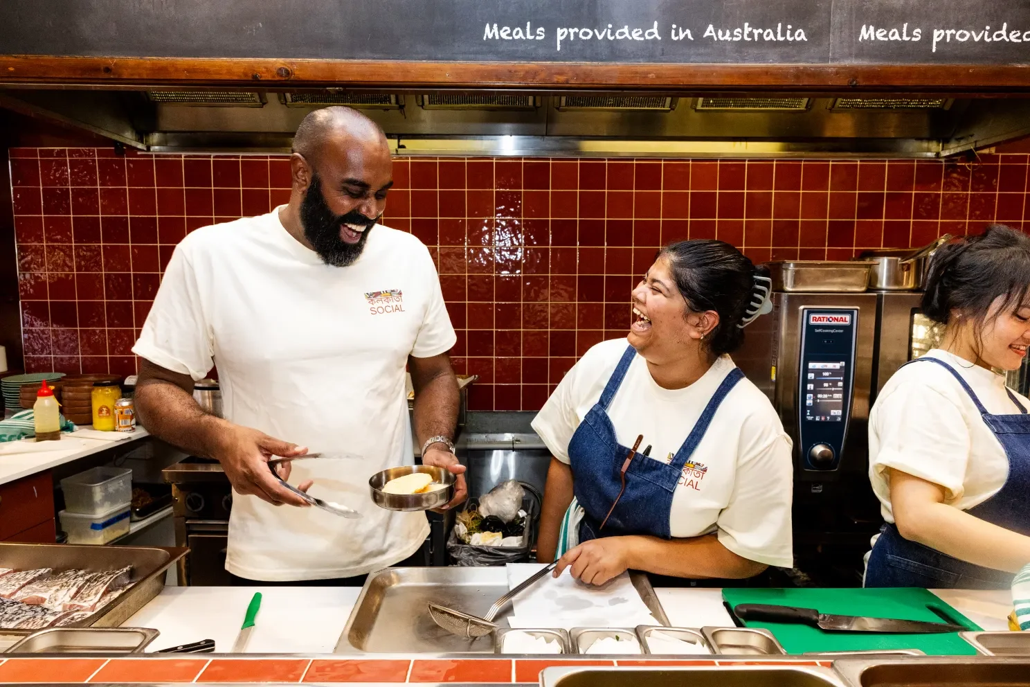 Two people in a commercial kitchen, laughing and sharing a moment. One man is handing a plate with food to a woman, both are wearing white shirts, and the woman is also wearing a blue apron.