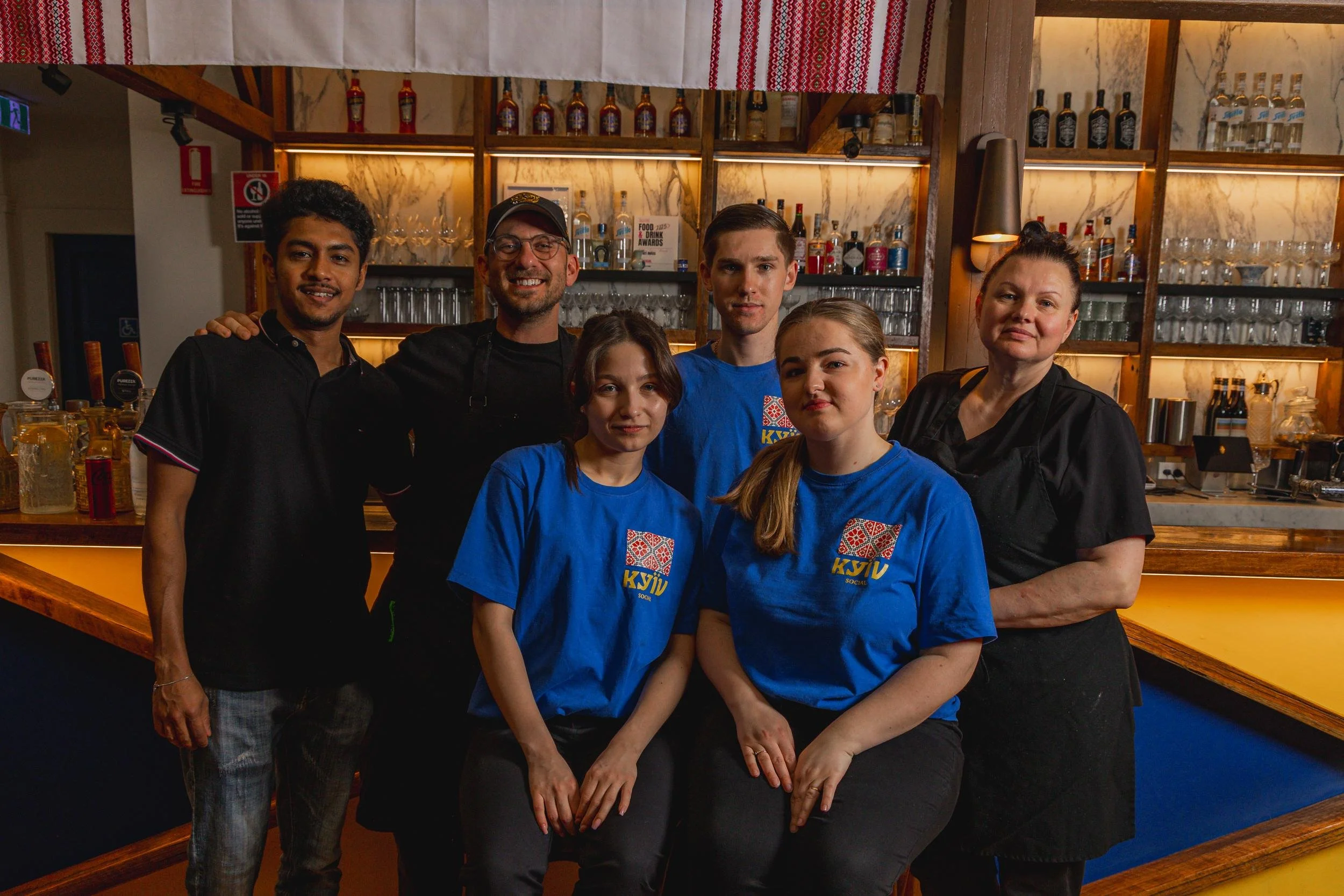 Group photo of six restaurant staff members, including chefs and waitstaff, standing together behind a bar in a restaurant.