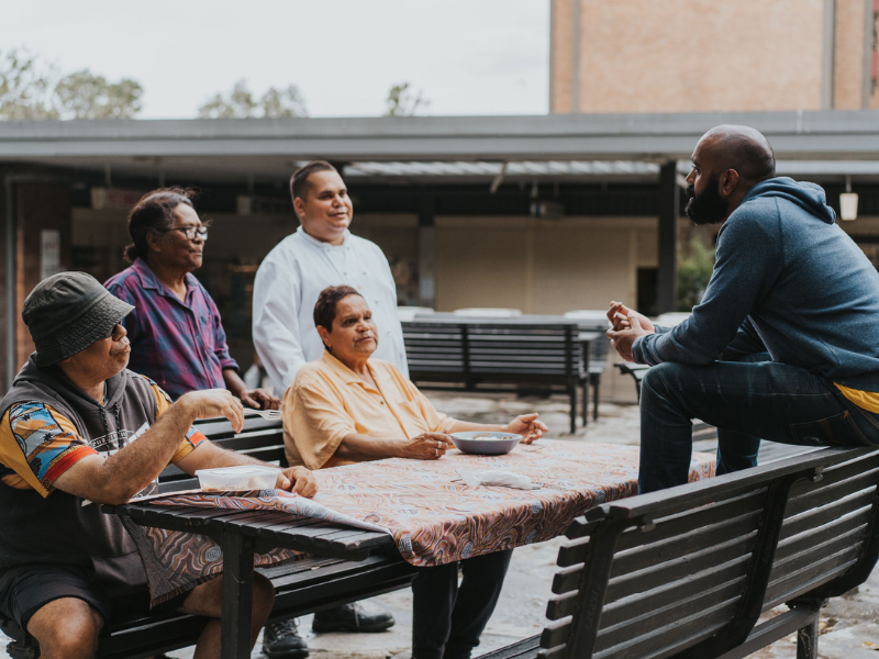 A group of five people gathered outdoors around a table, engaged in conversation with a man sitting on a bench. The group includes four women, one of whom is seated, and one standing man, wearing casual clothing including a hoodie and glasses. The sc