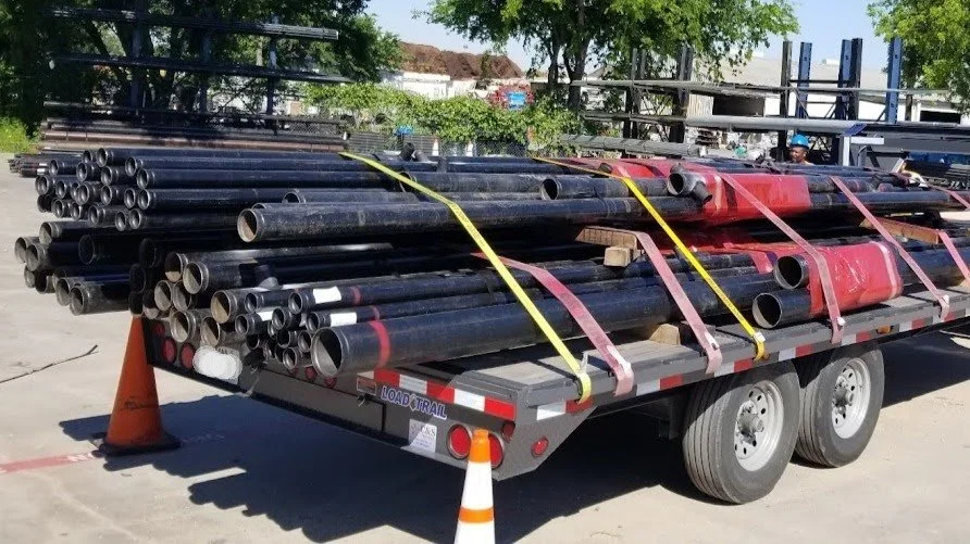 A flatbed truck loaded with black metal pipes secured with yellow and pink straps, with orange traffic cones around it, outdoors in a parking lot with trees and industrial background.