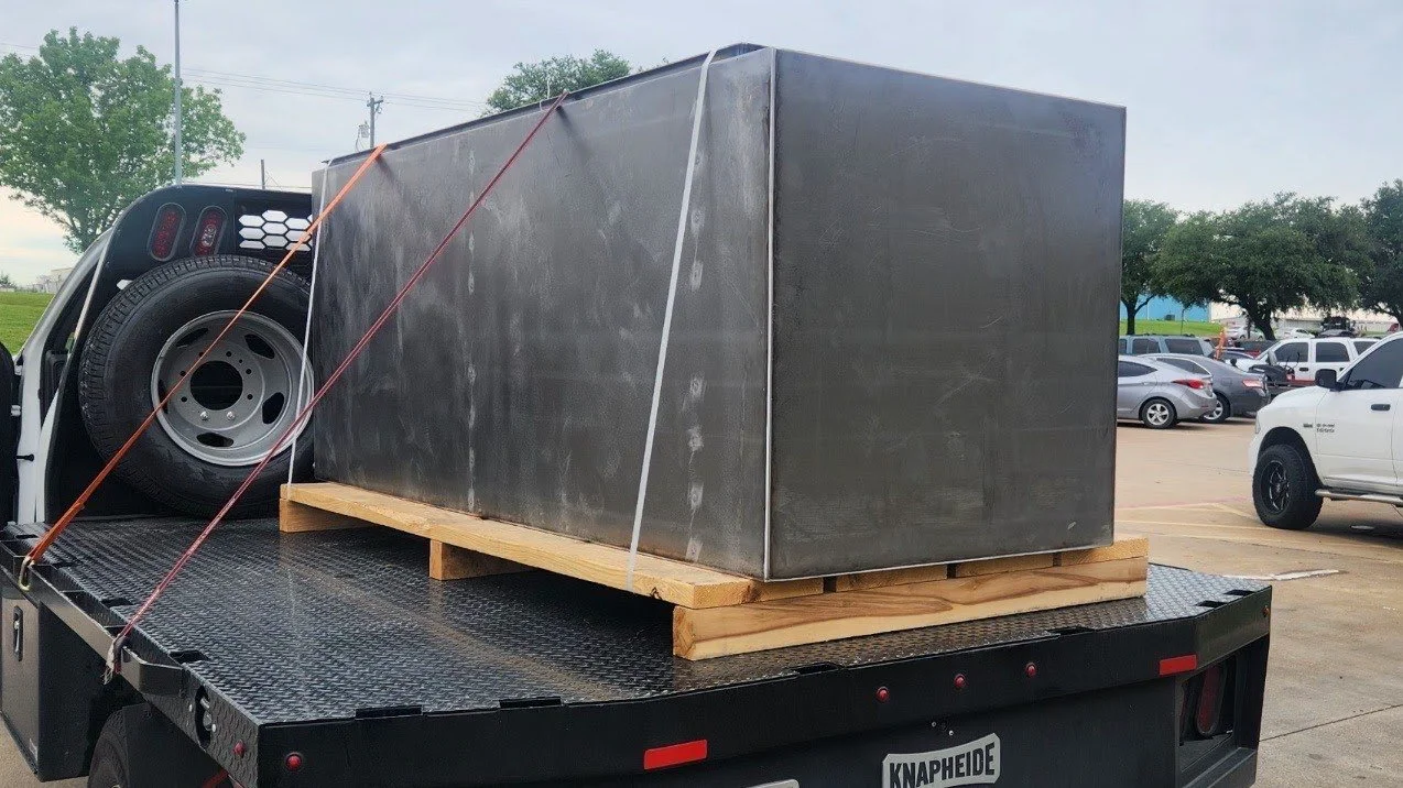 A large metal box secured on a wooden pallet with straps on a flatbed truck, parked in a parking lot.