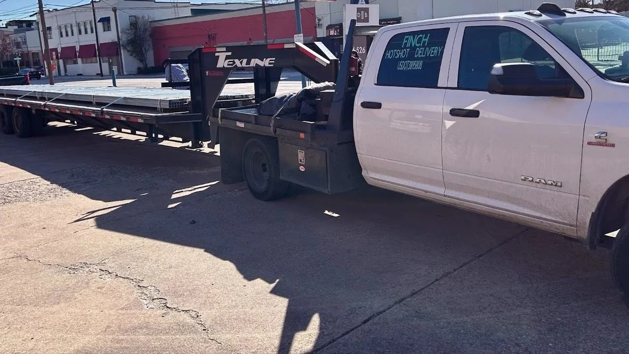 A white RAM pickup truck with a flatbed trailer attached, parked on a city street. The truck has signage on the window advertising hotshot delivery for Finch.